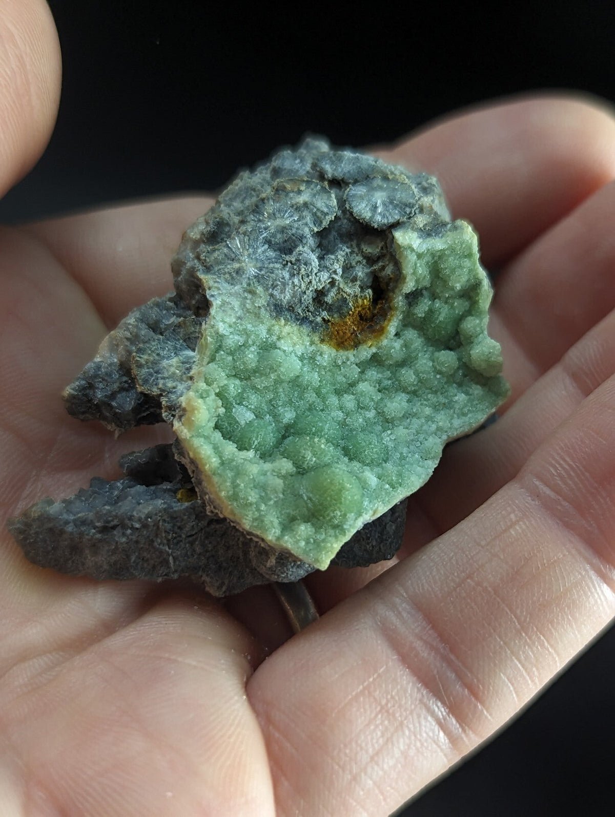 A hand holds a rough, irregularly shaped mineral specimen featuring vibrant green crystalline formations contrasted with dark gray matrix and a small patch of yellowish-orange material.