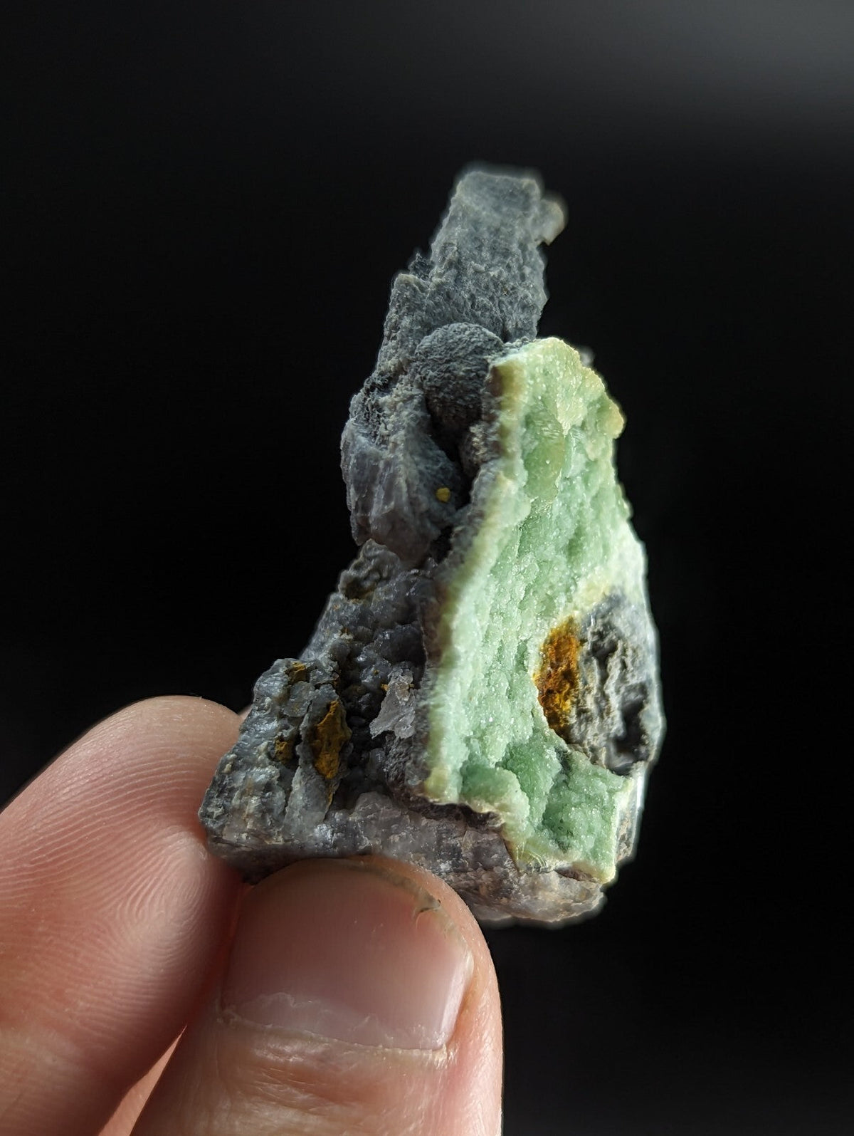 A hand holds a rough, irregularly shaped mineral specimen featuring a prominent light green crystalline section with subtle yellowish inclusions and dark gray matrix material.