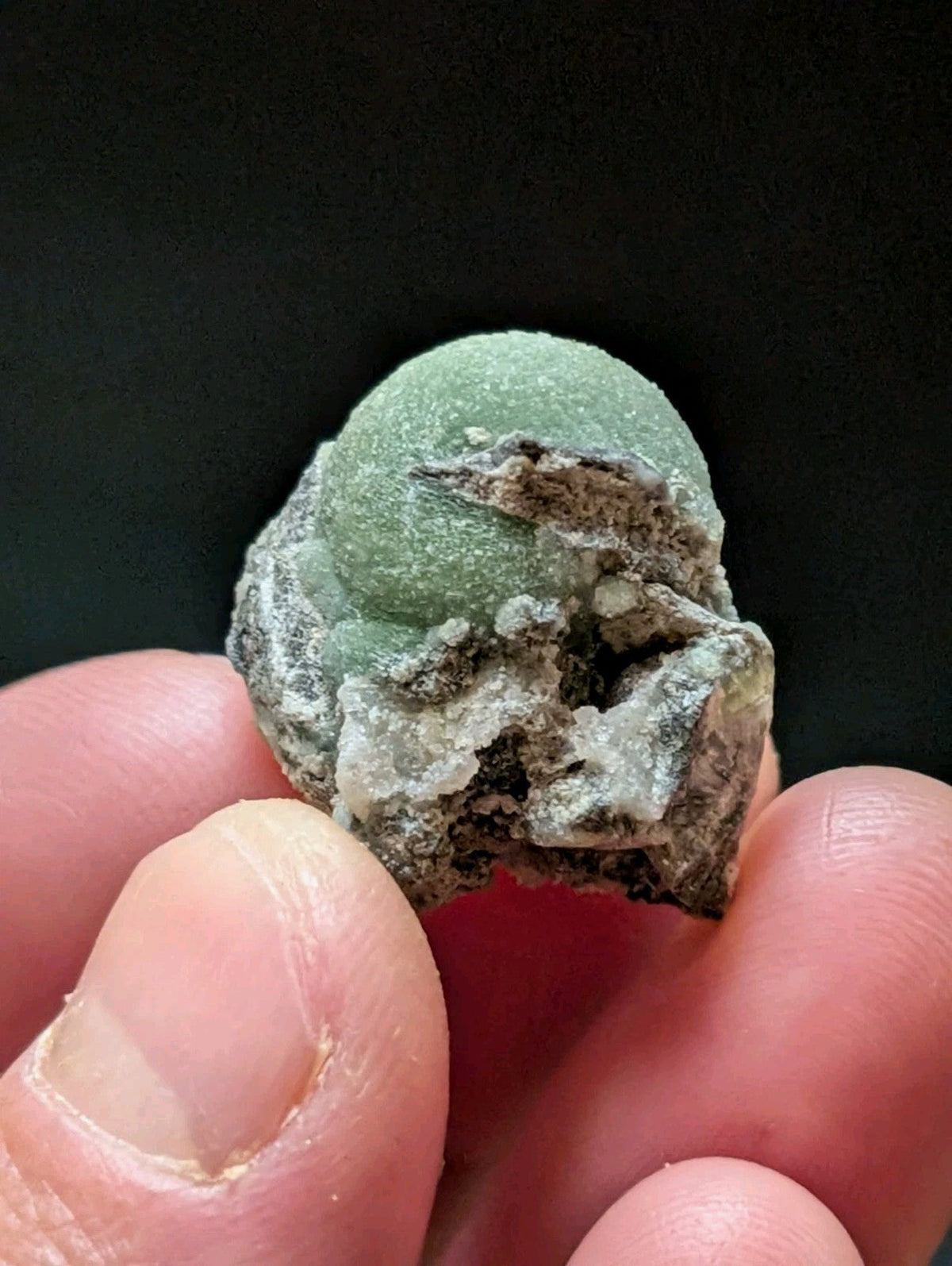 A smooth, pale green spherical mineral specimen with a rough, earthy matrix surrounding it.