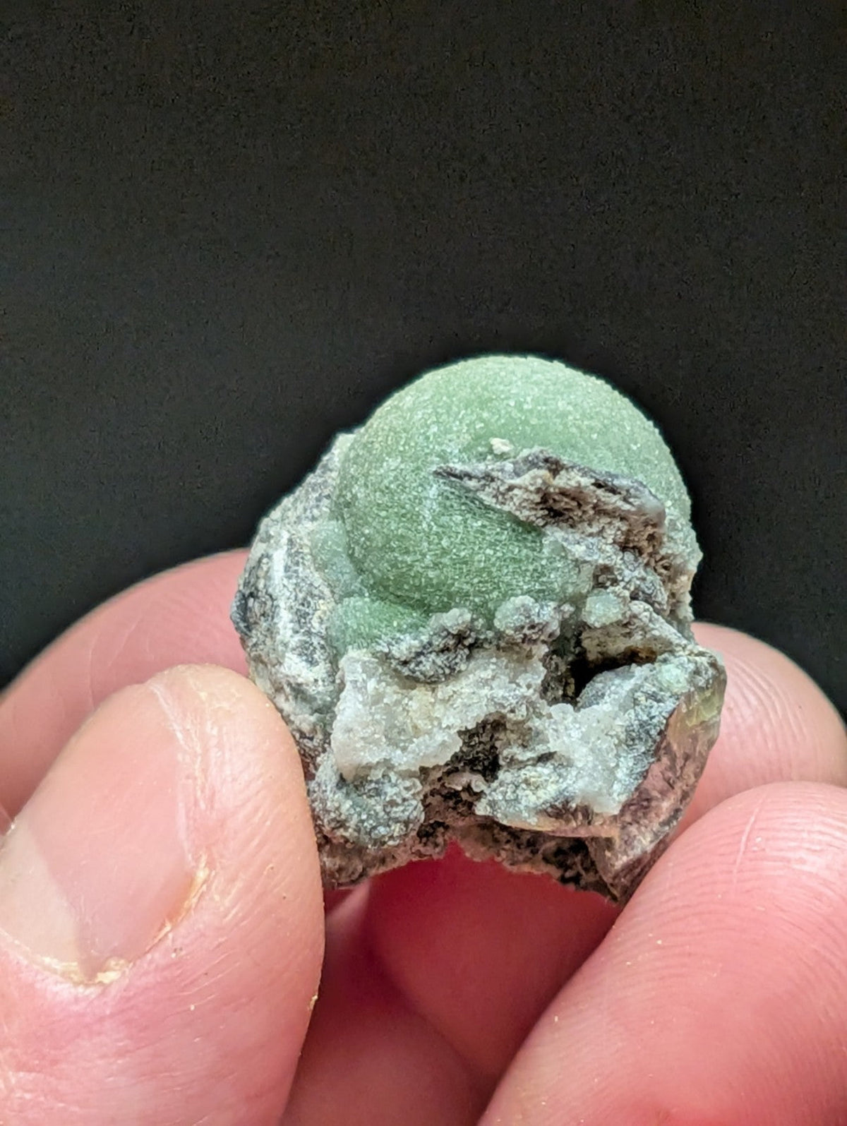 A green, rounded mineral specimen with a rough, textured surface and patches of darker material.