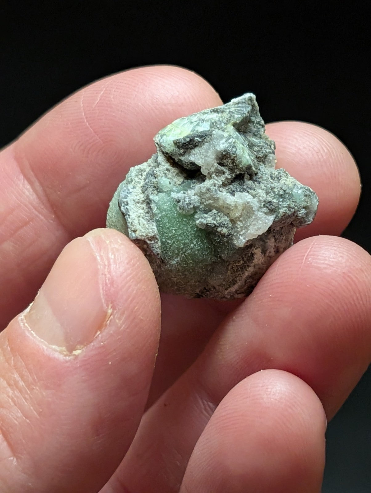 A rough, irregularly shaped mineral specimen with a mottled green and white surface, held gently between fingers.
