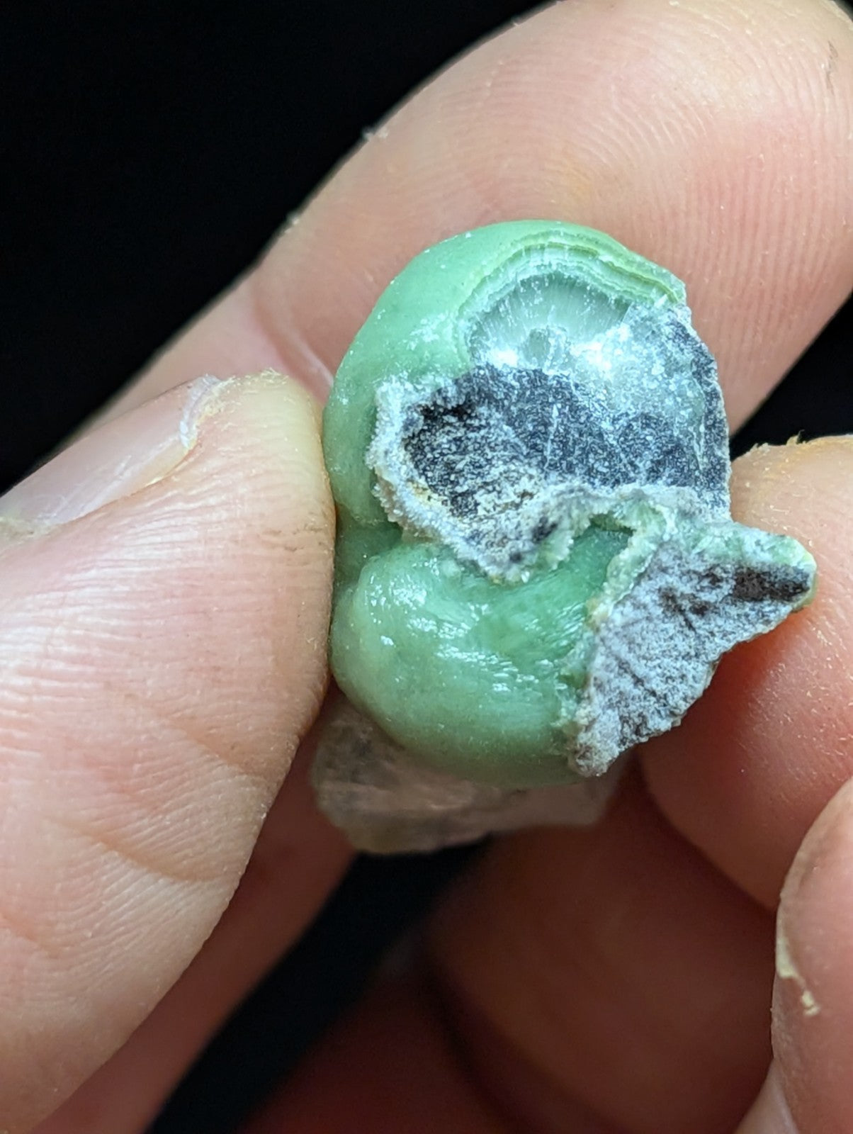 A green, irregularly shaped mineral specimen with a partially broken surface revealing darker interior layers and white crystalline deposits.