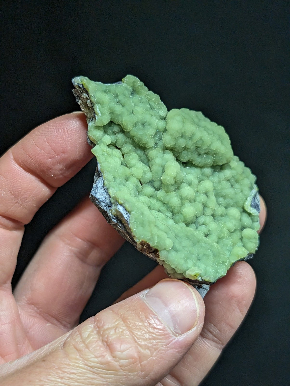 A hand holds a vibrant green, bumpy mineral specimen with a rough, textured surface and some darker grayish edges.
