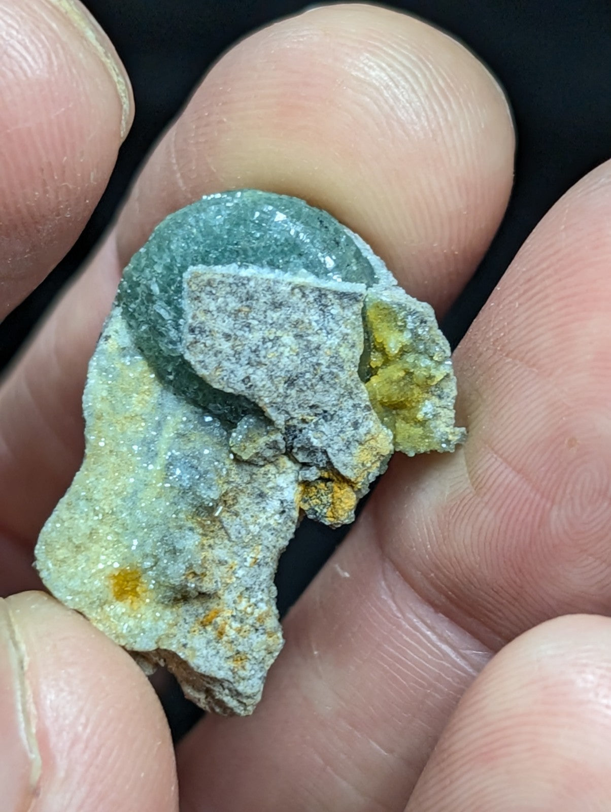 A hand holds a small, irregularly shaped mineral specimen featuring patches of greenish-blue and yellow-orange hues with a rough, crystalline texture.