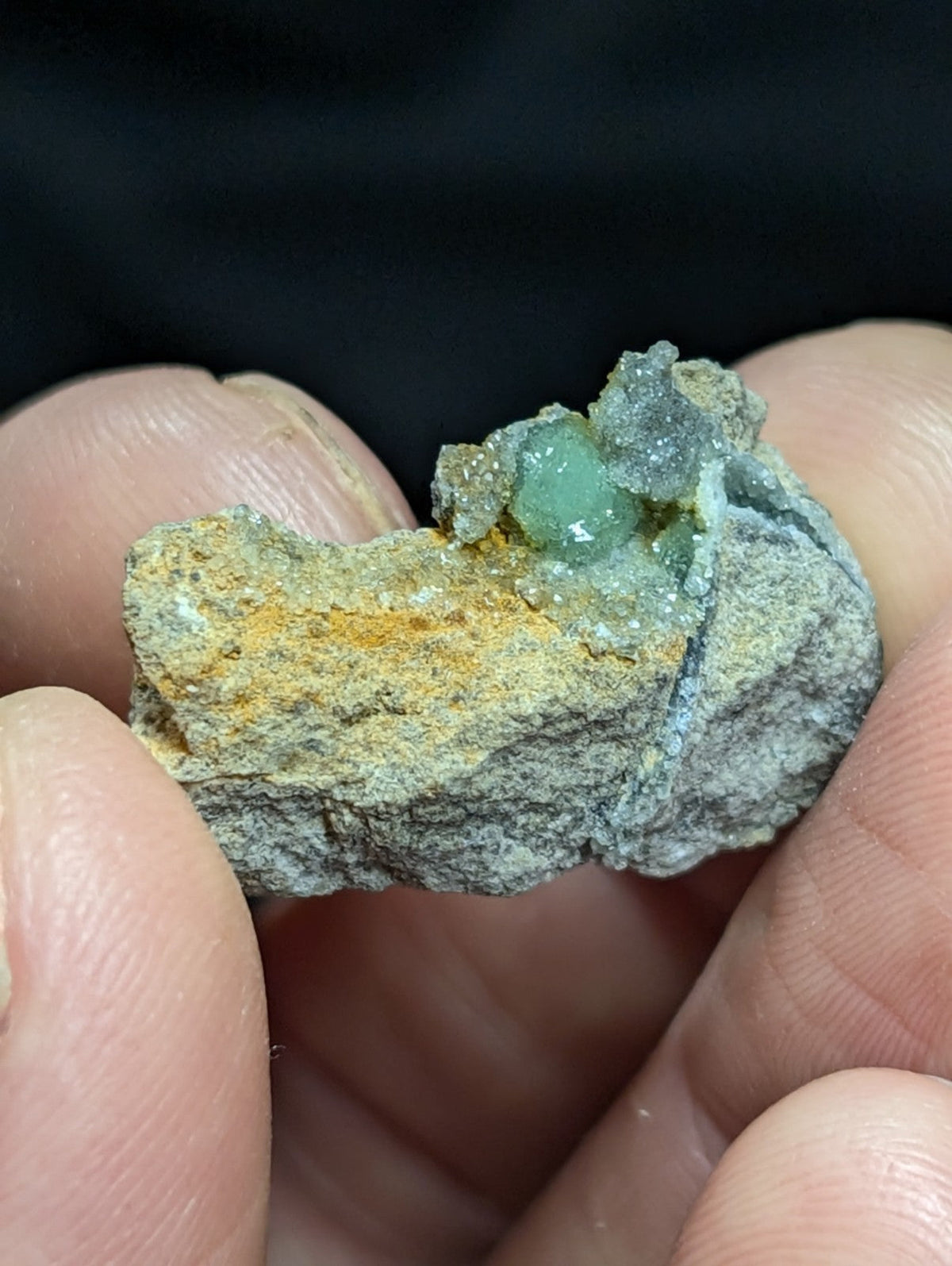 A hand holds a rough, grayish rock with patches of yellow mineral and small translucent green crystals growing from its surface.