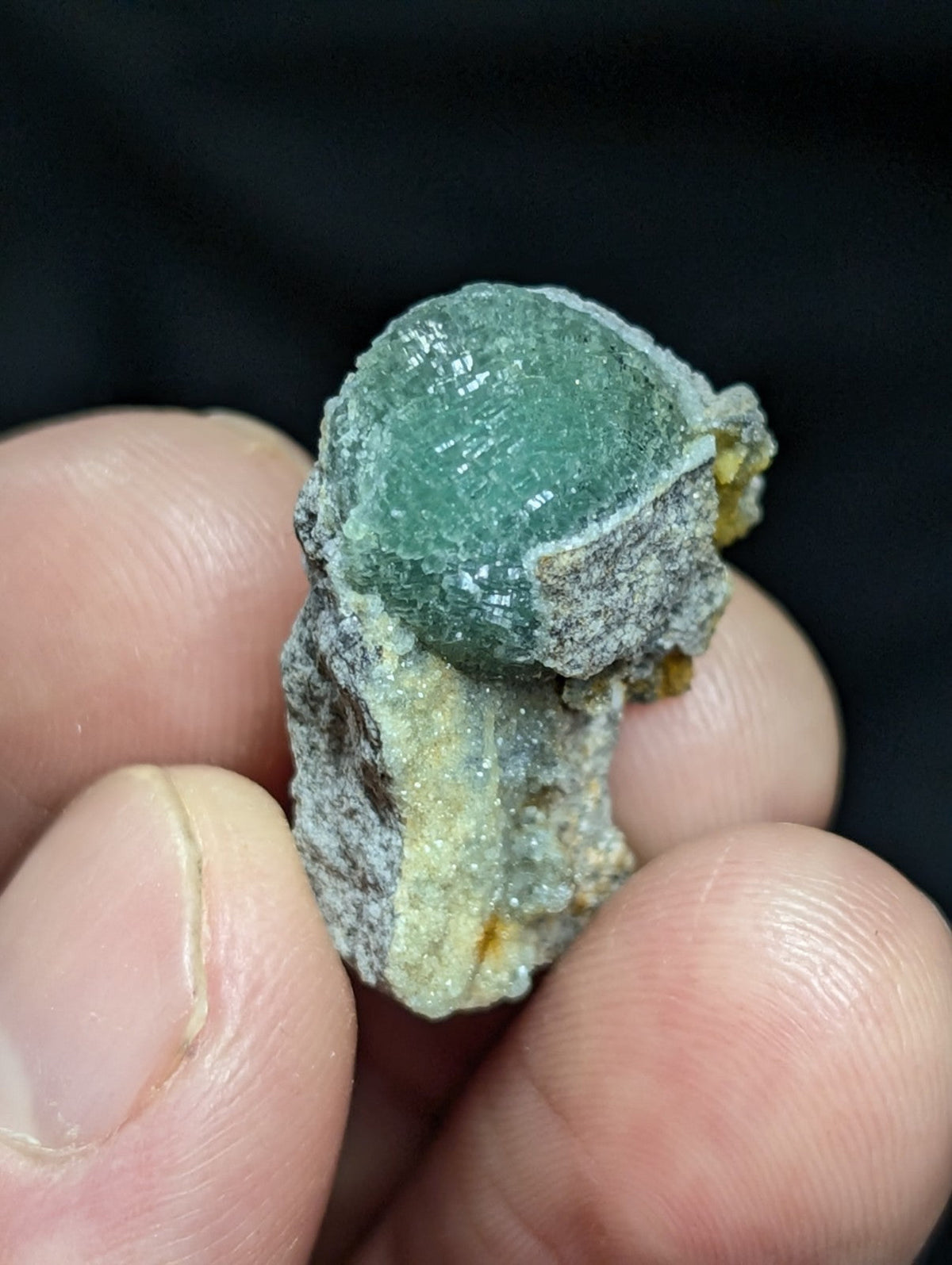 A translucent green spherical mineral specimen nestled in a rough, grayish matrix with hints of yellow and white crystalline material.