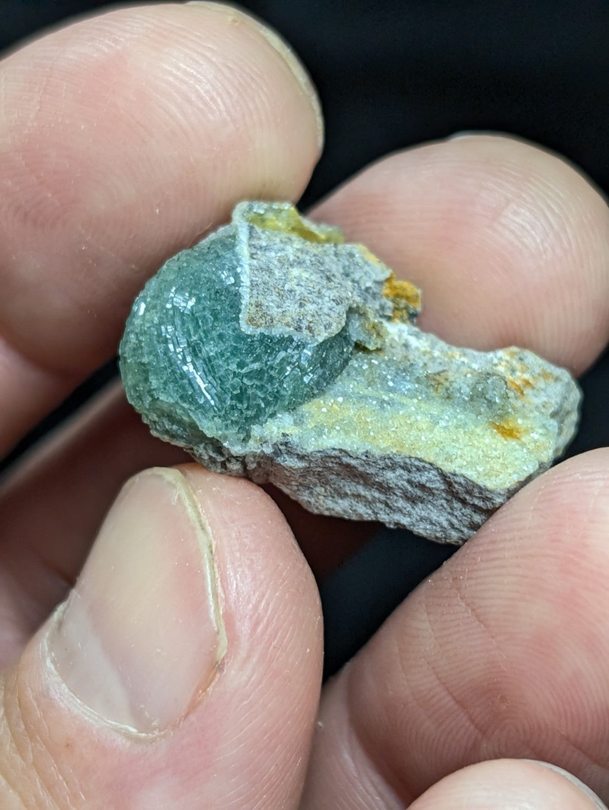 A hand holds a rough, irregularly shaped mineral specimen featuring translucent green crystals with patches of grayish matrix and small yellow inclusions.