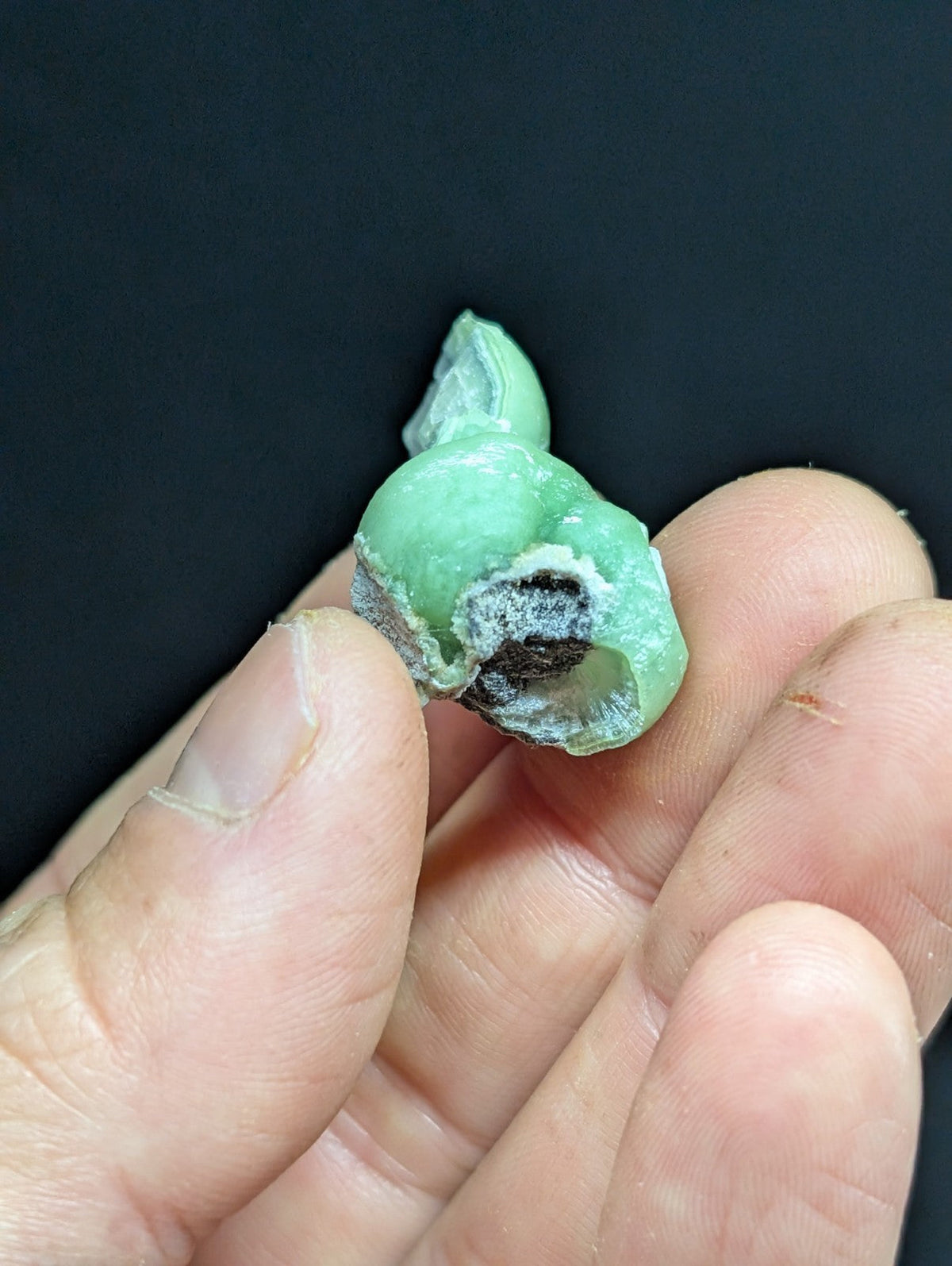 A hand holds a small, irregularly shaped green gemstone with a rough, natural surface and visible internal dark inclusions.