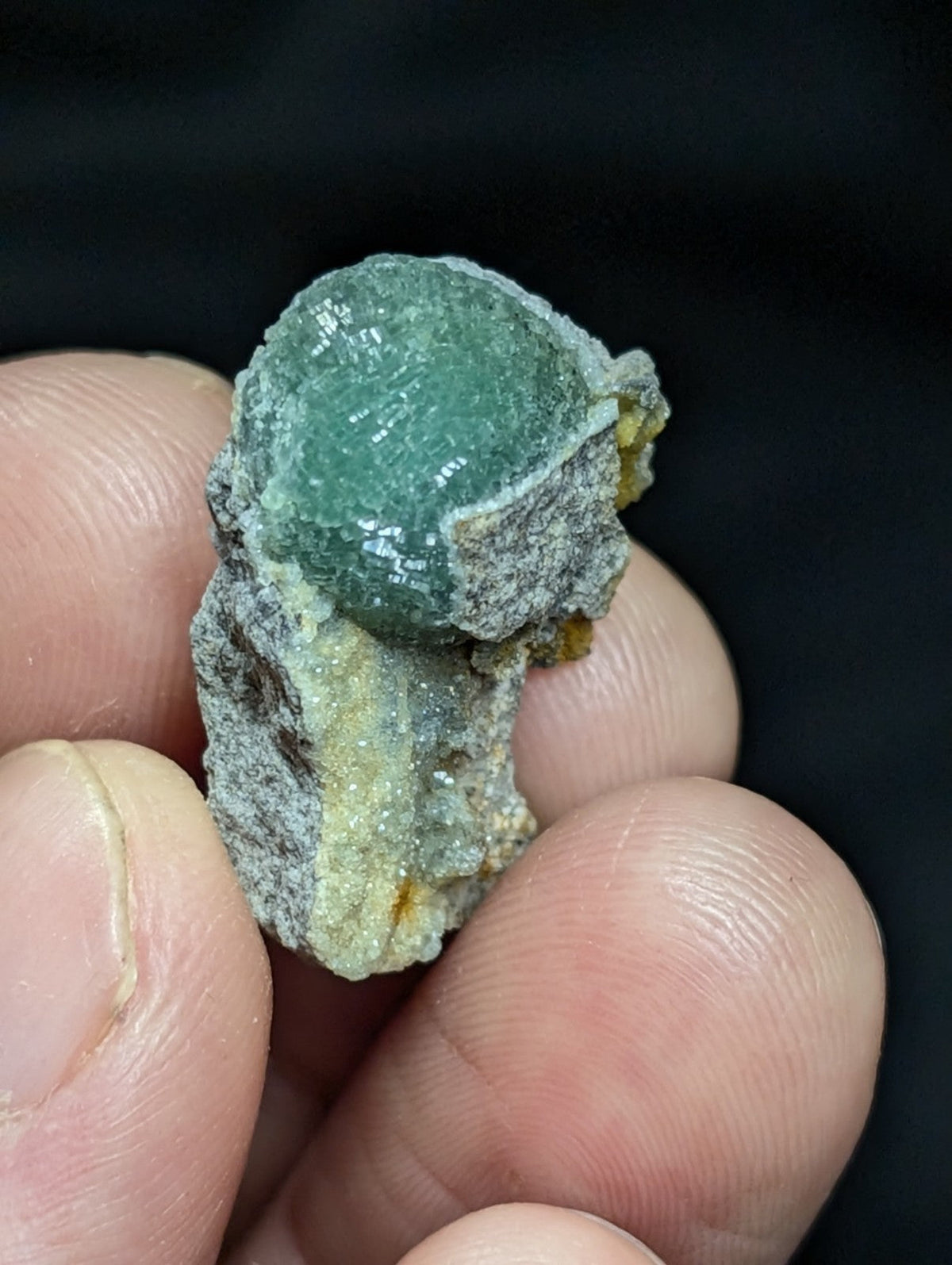 A translucent green gemstone nestled in a rough, grayish matrix with hints of yellow.