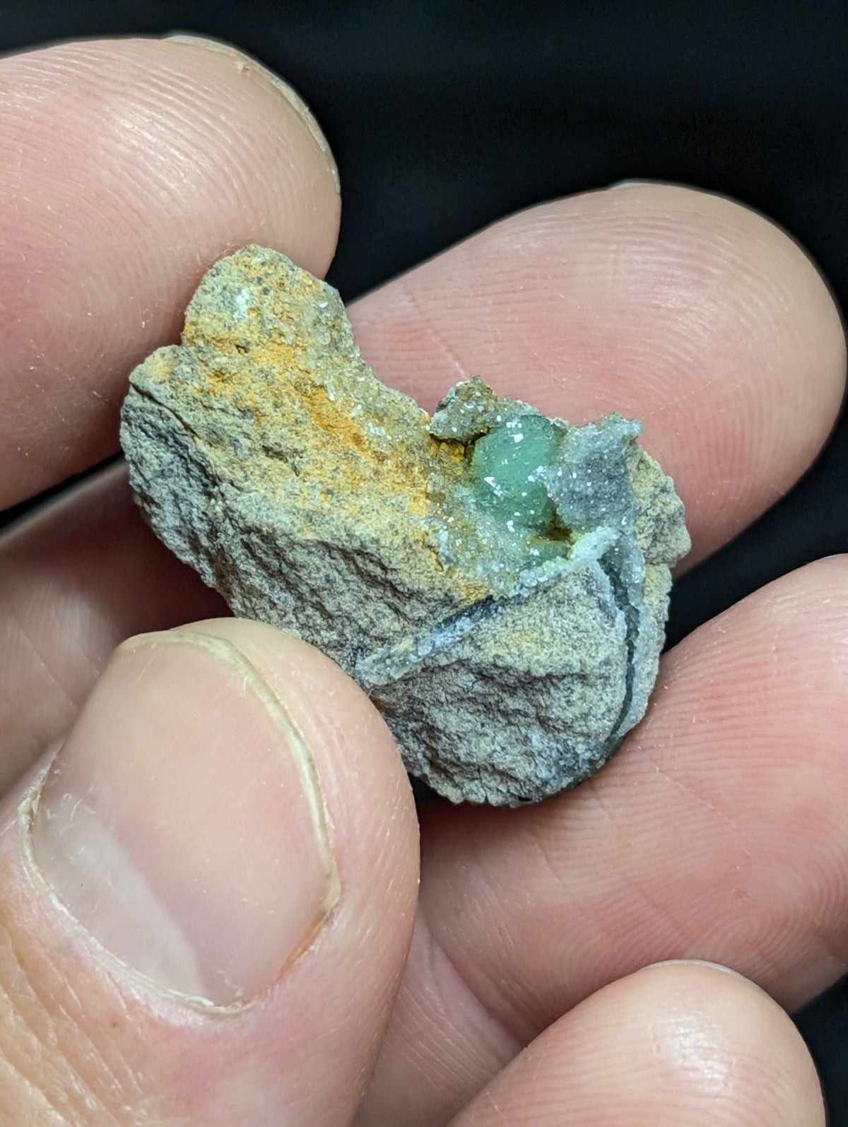 A small, irregularly shaped mineral specimen featuring a cluster of translucent green crystals nestled within a rough-textured matrix of gray and yellowish rock.
