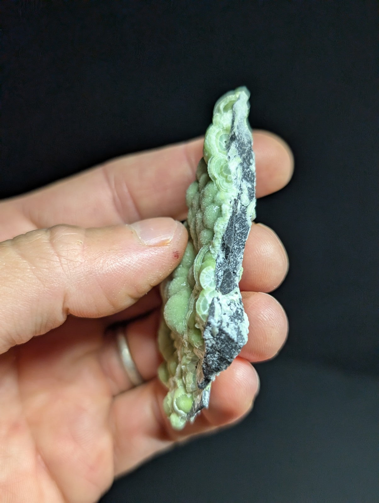 A hand holds a carved jadeite piece with intricate, bead-like patterns in translucent green and dark gray veining.