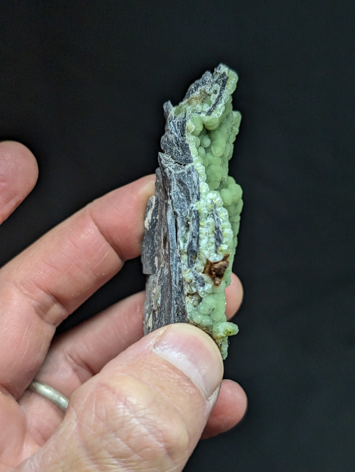 A hand holds a striking green mineral specimen with textured, bumpy growths against a dark gray matrix.