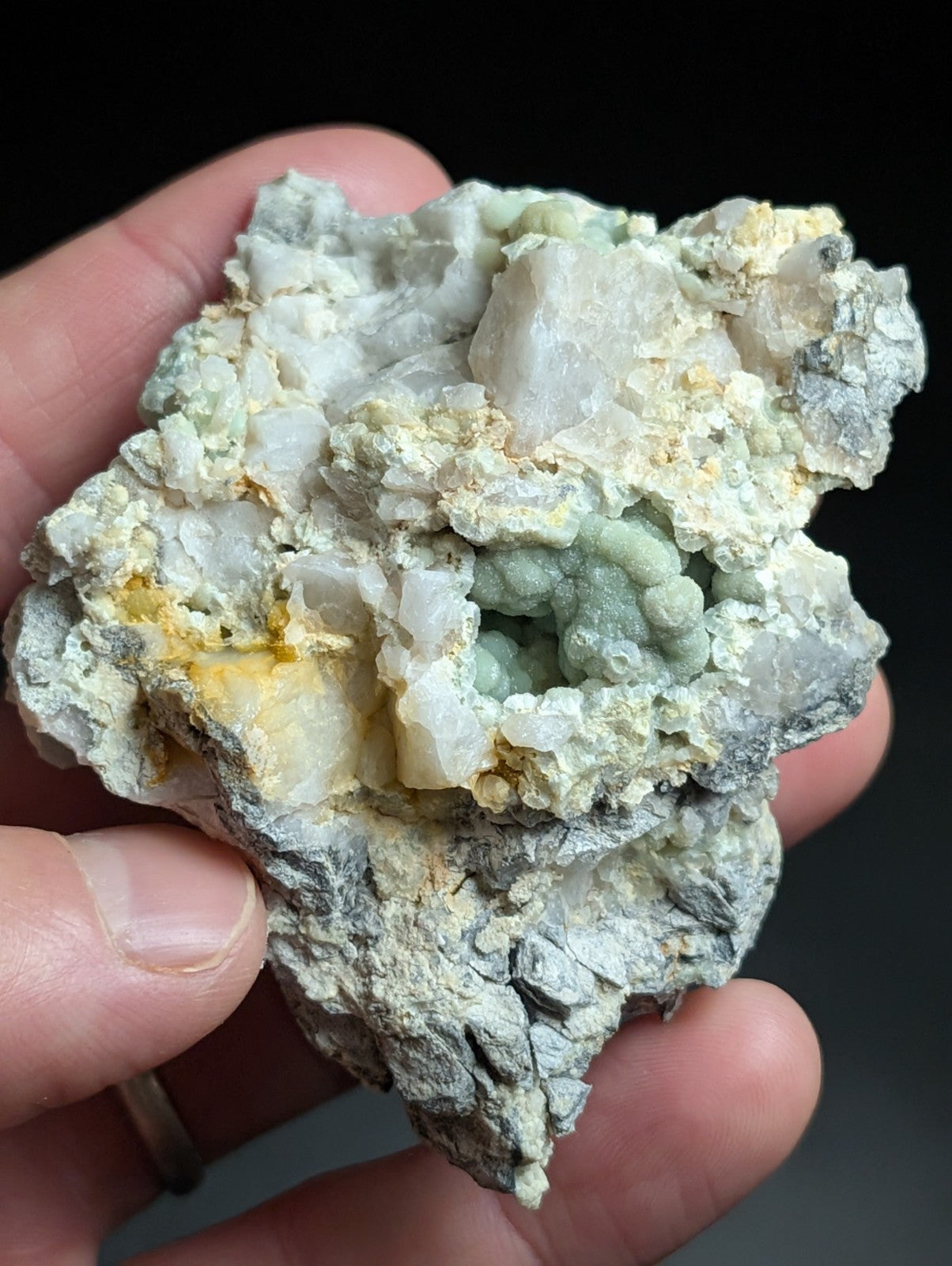 A hand holds a rough, irregularly shaped mineral specimen featuring clusters of translucent white and pale green crystals interspersed with patches of yellowish material.