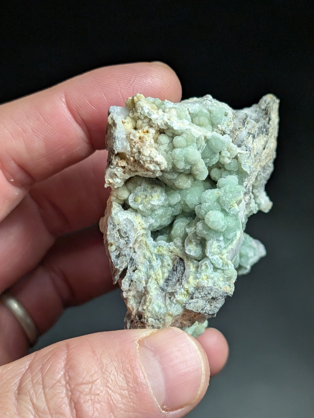 A hand holds a rough, irregularly shaped mineral specimen featuring clusters of pale green crystals growing from a darker gray matrix.