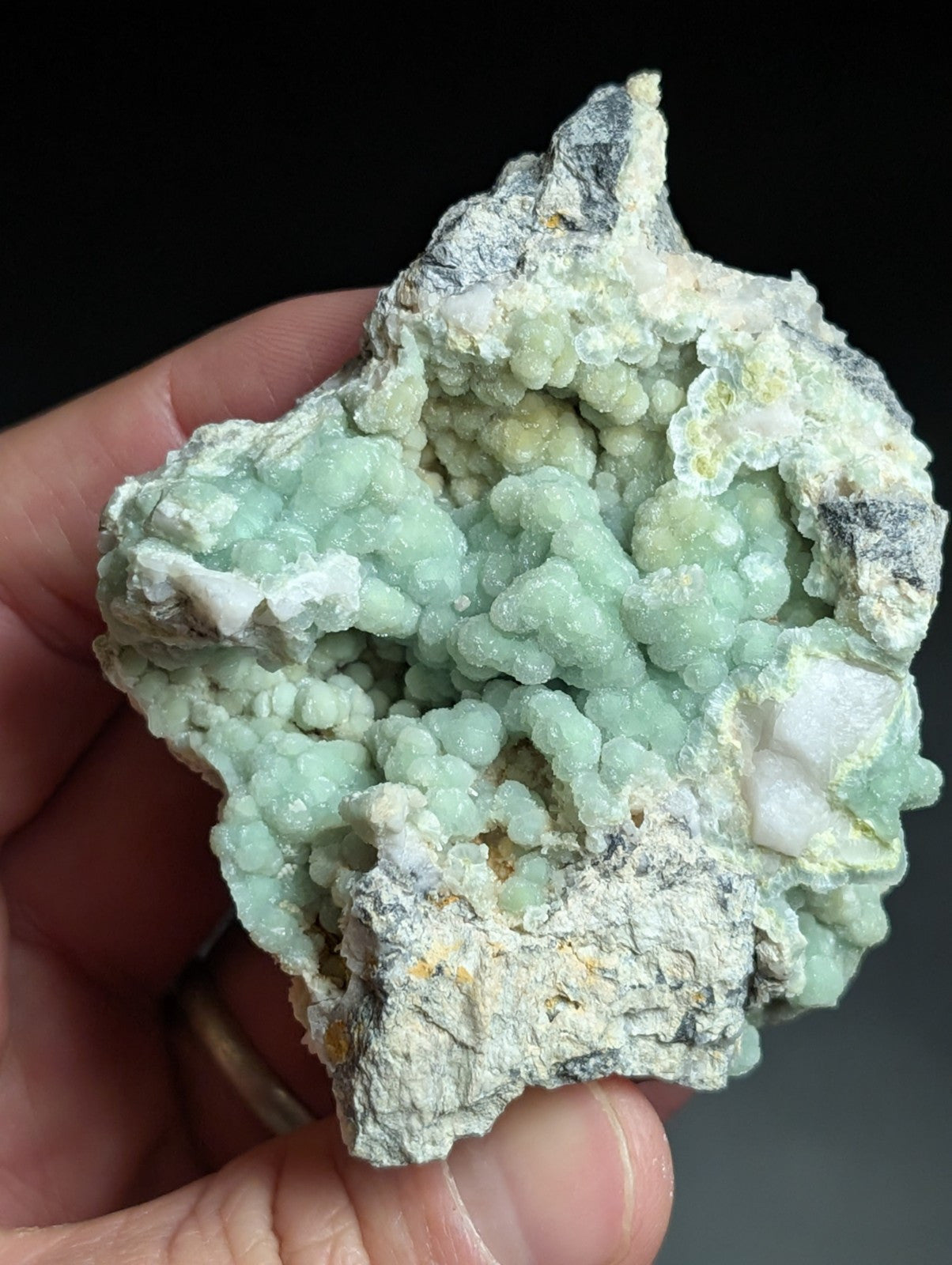 A hand holds a natural mineral specimen featuring clusters of pale green, bubbly crystals growing from a rougher gray and white matrix.