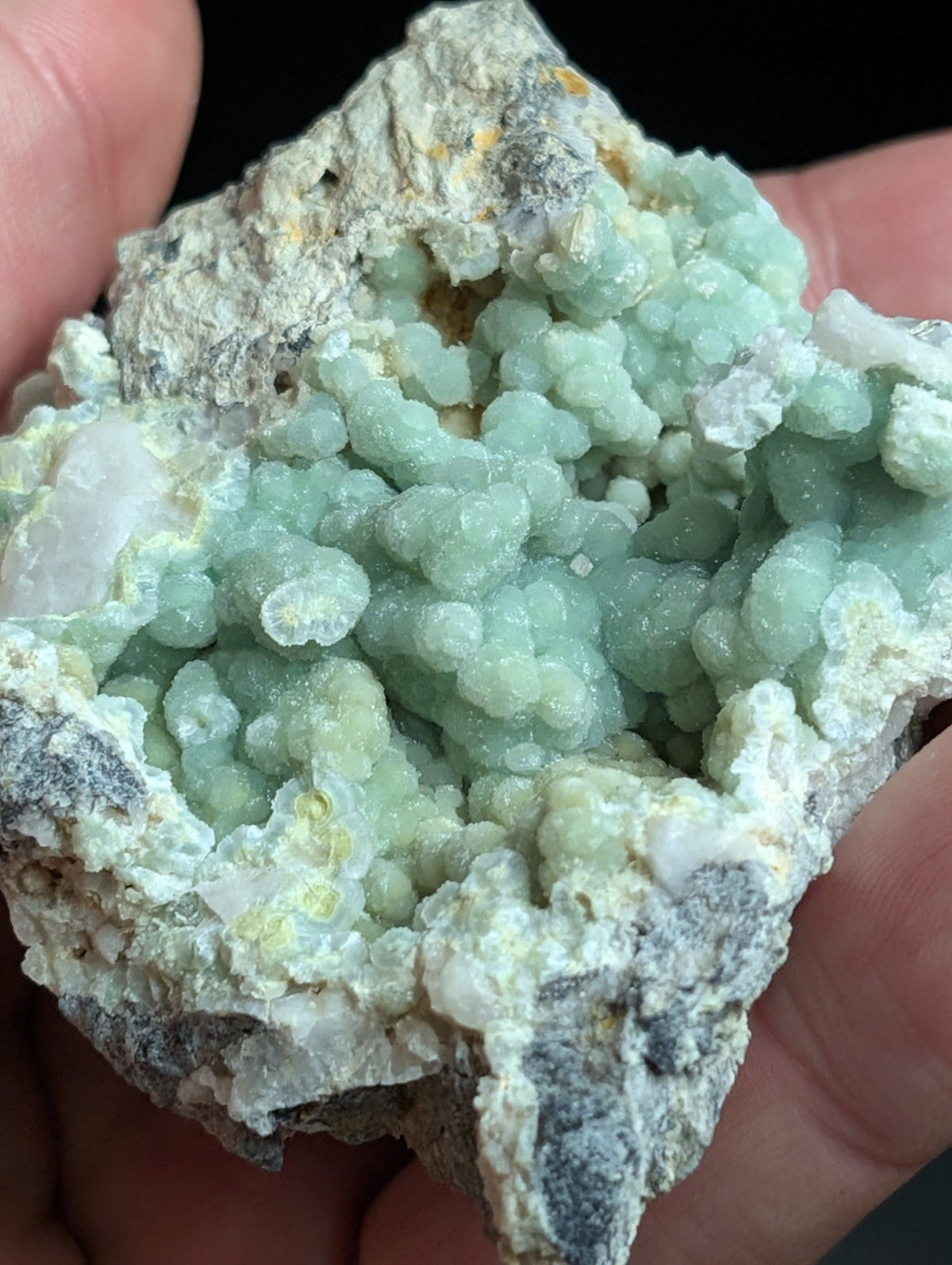 A hand holds a natural mineral specimen featuring clusters of translucent, light green crystals growing from a rough, grayish matrix.