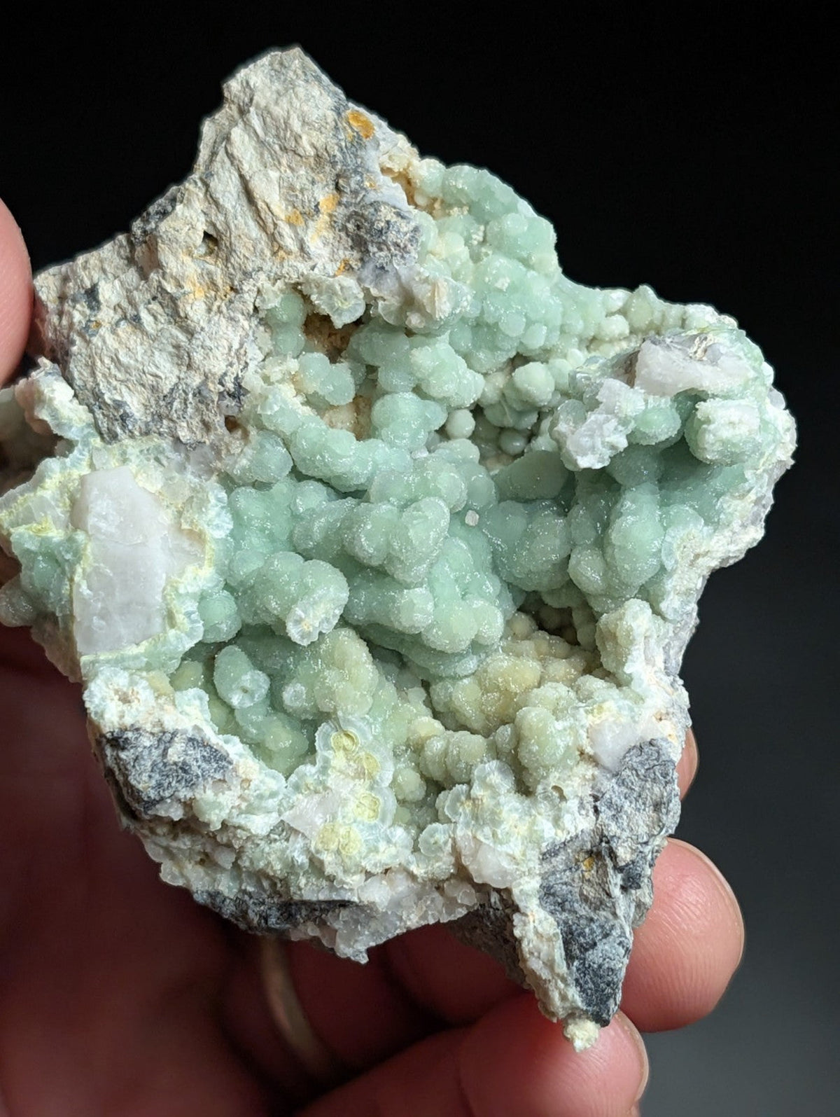 A hand holds a natural mineral specimen featuring clusters of pale green, rounded crystals growing from a rougher gray and white rock matrix.