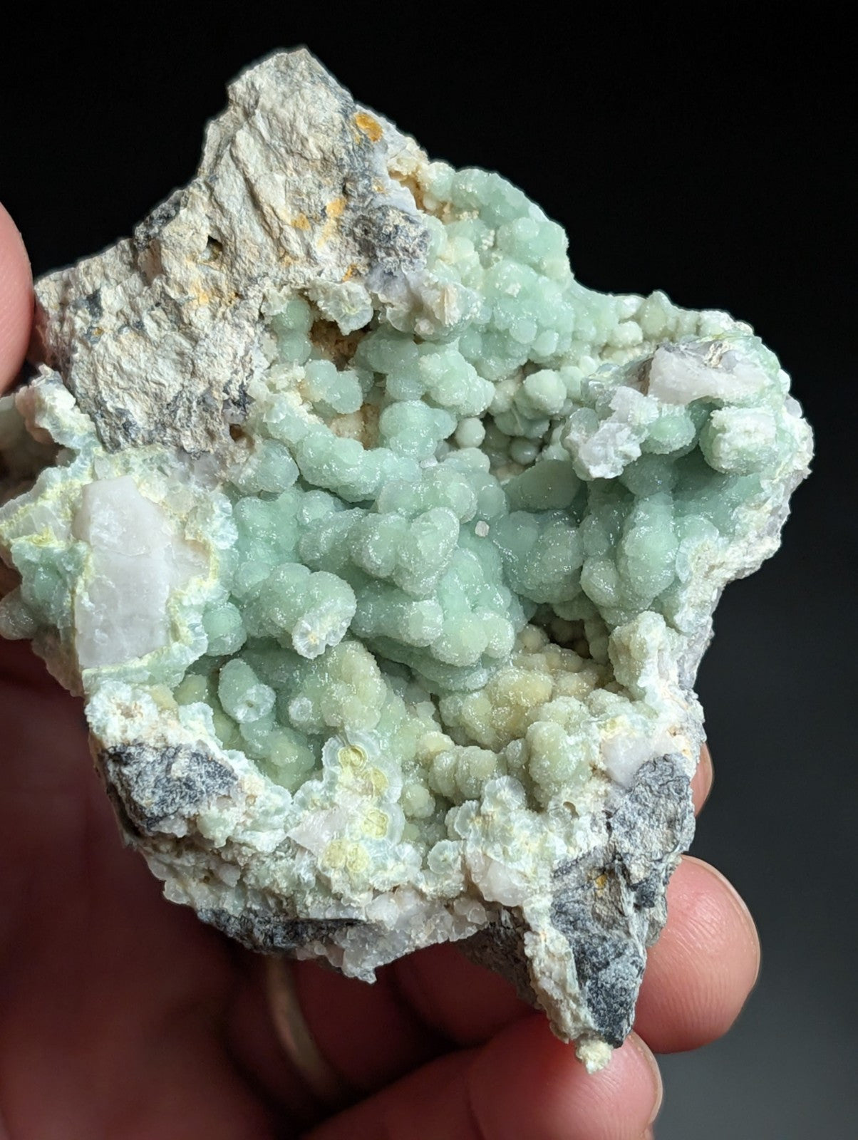 A hand holds a natural mineral specimen featuring clusters of pale green, rounded crystals growing from a rougher gray and white rock matrix.