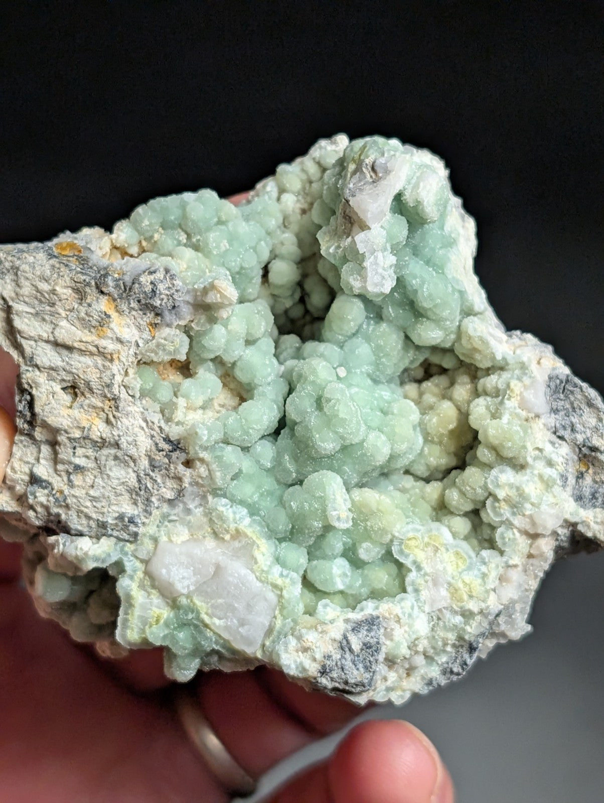 A hand holds a striking greenish-blue mineral specimen with clustered, bulbous crystals growing from a rougher gray matrix.