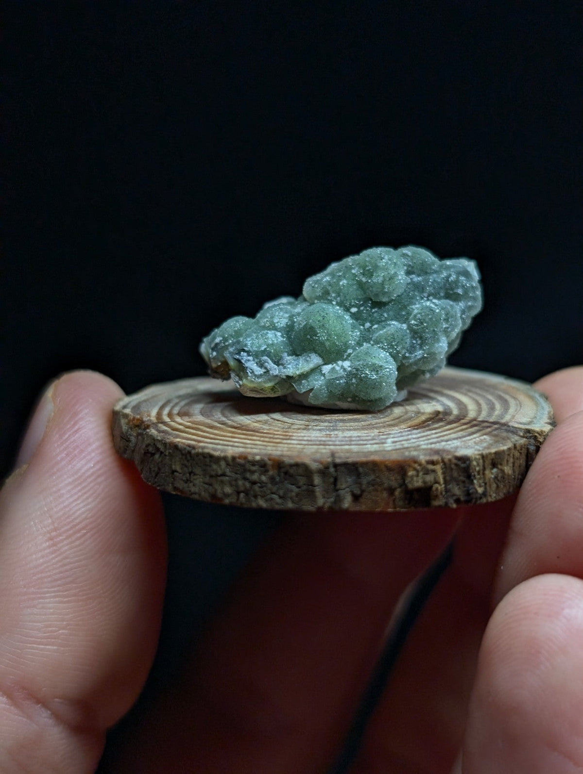 A delicate, translucent green mineral specimen with a clustered, crystalline texture rests on a rustic wooden slice.