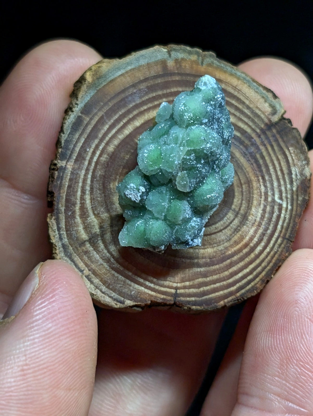 A cluster of translucent green crystals with a slightly frosted, glassy surface rests on a circular wooden slice.
