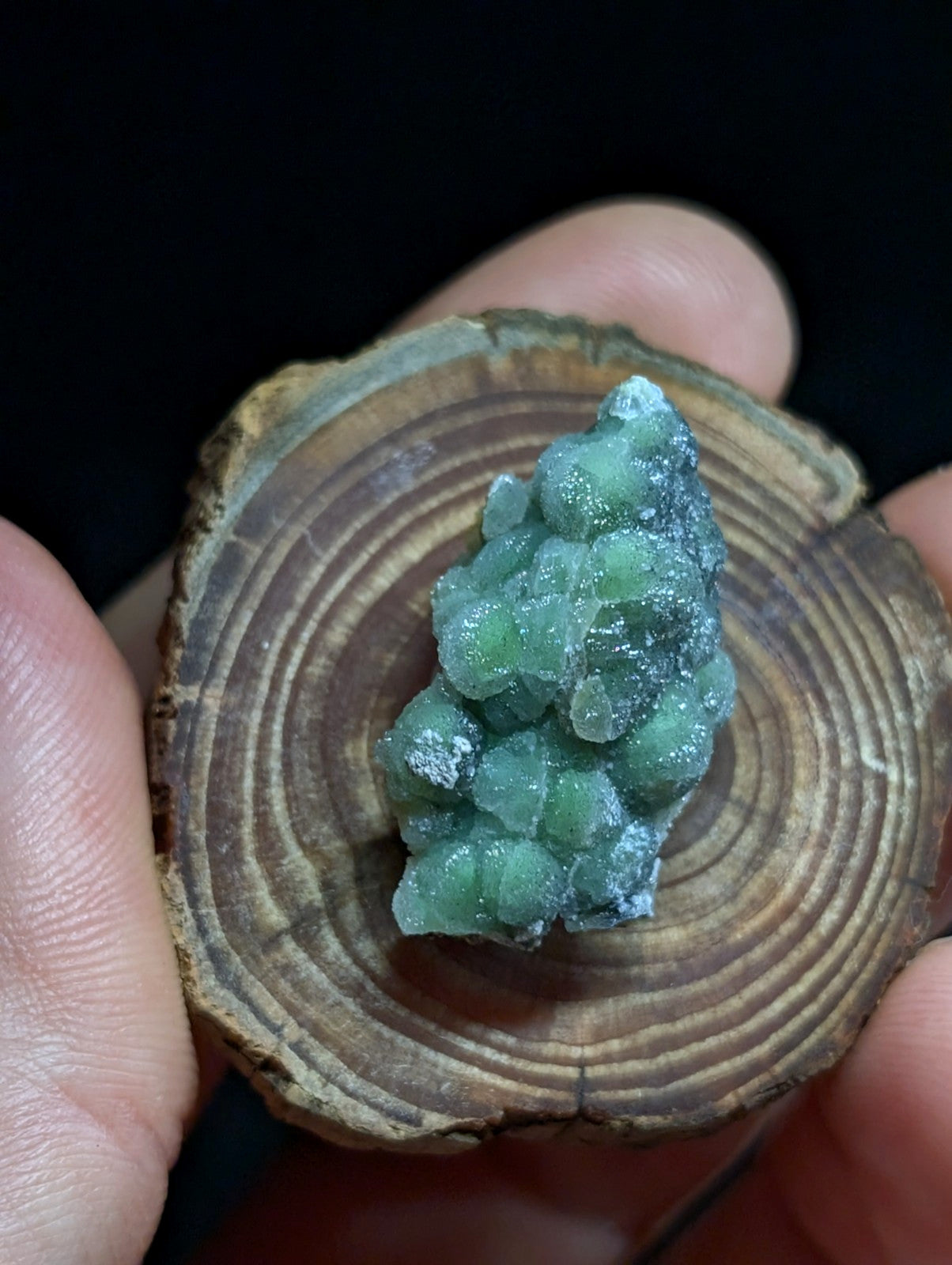 A cluster of translucent green crystals with a slightly frosted, crystalline texture rests on a circular wooden slice.