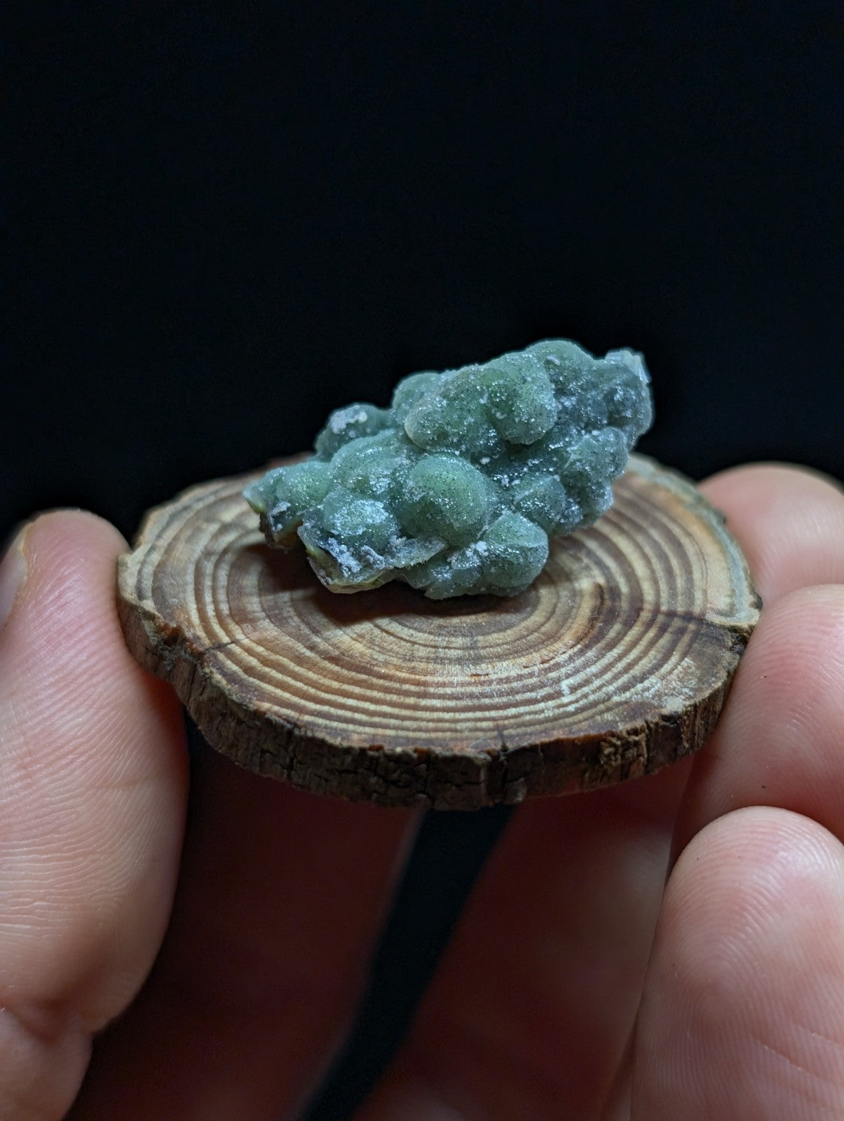 A cluster of translucent green mineral crystals with a rough, bumpy texture rests on a circular wooden slice.