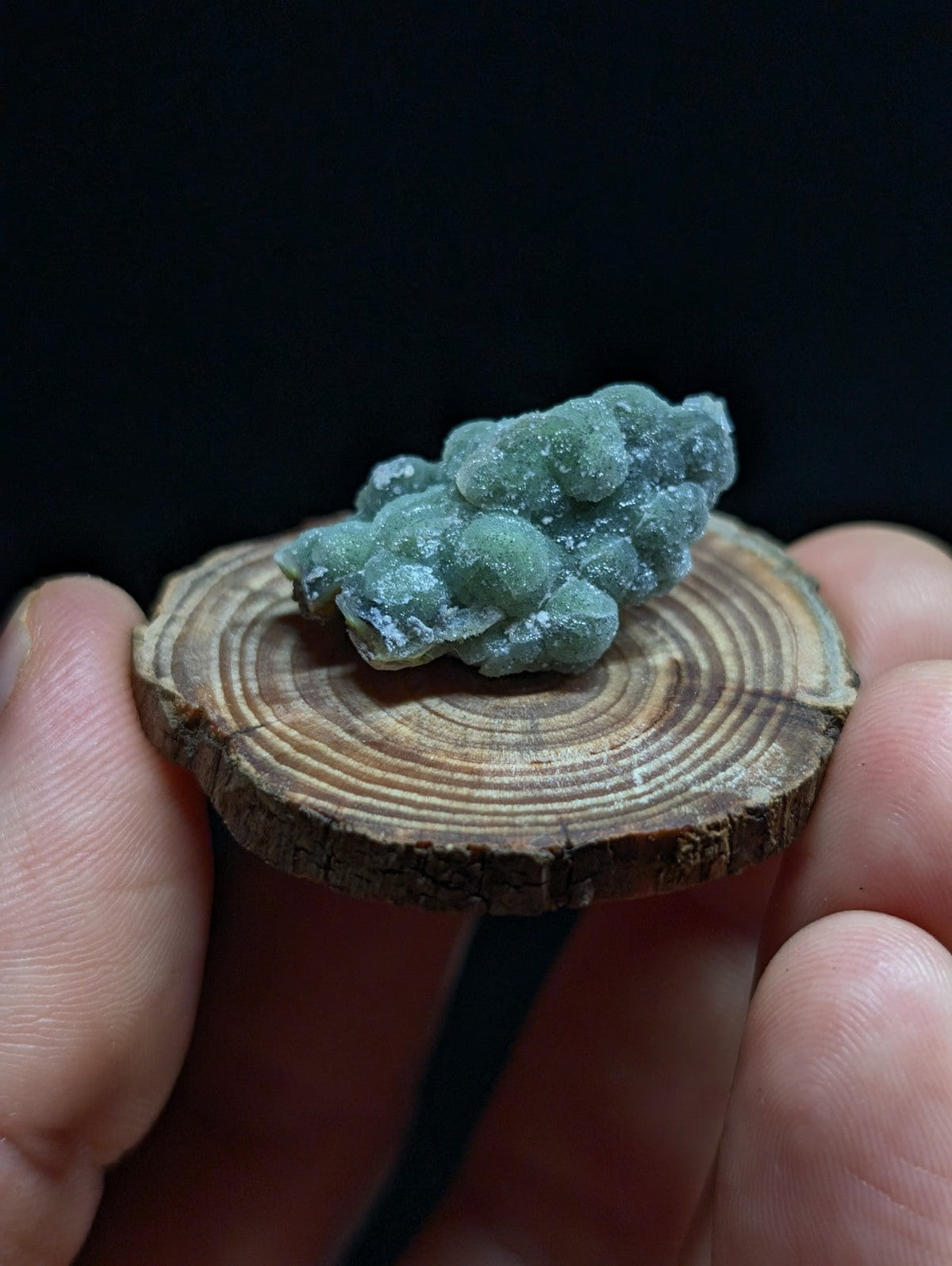A cluster of translucent green mineral crystals with a rough, bumpy texture rests on a circular wooden slice.