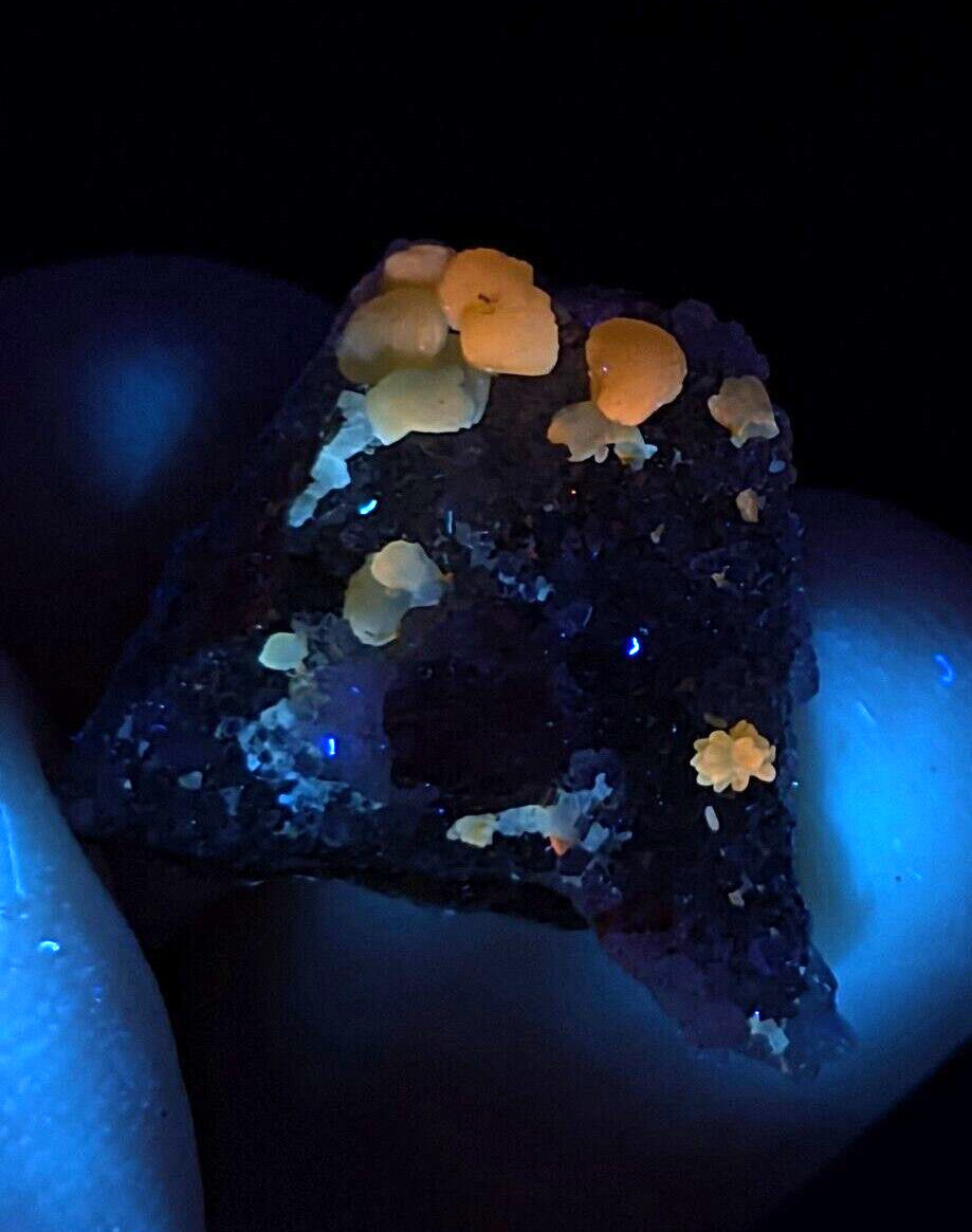 A glowing mineral specimen featuring vibrant orange and pale yellow fluorescent crystals growing from a dark, textured base.
