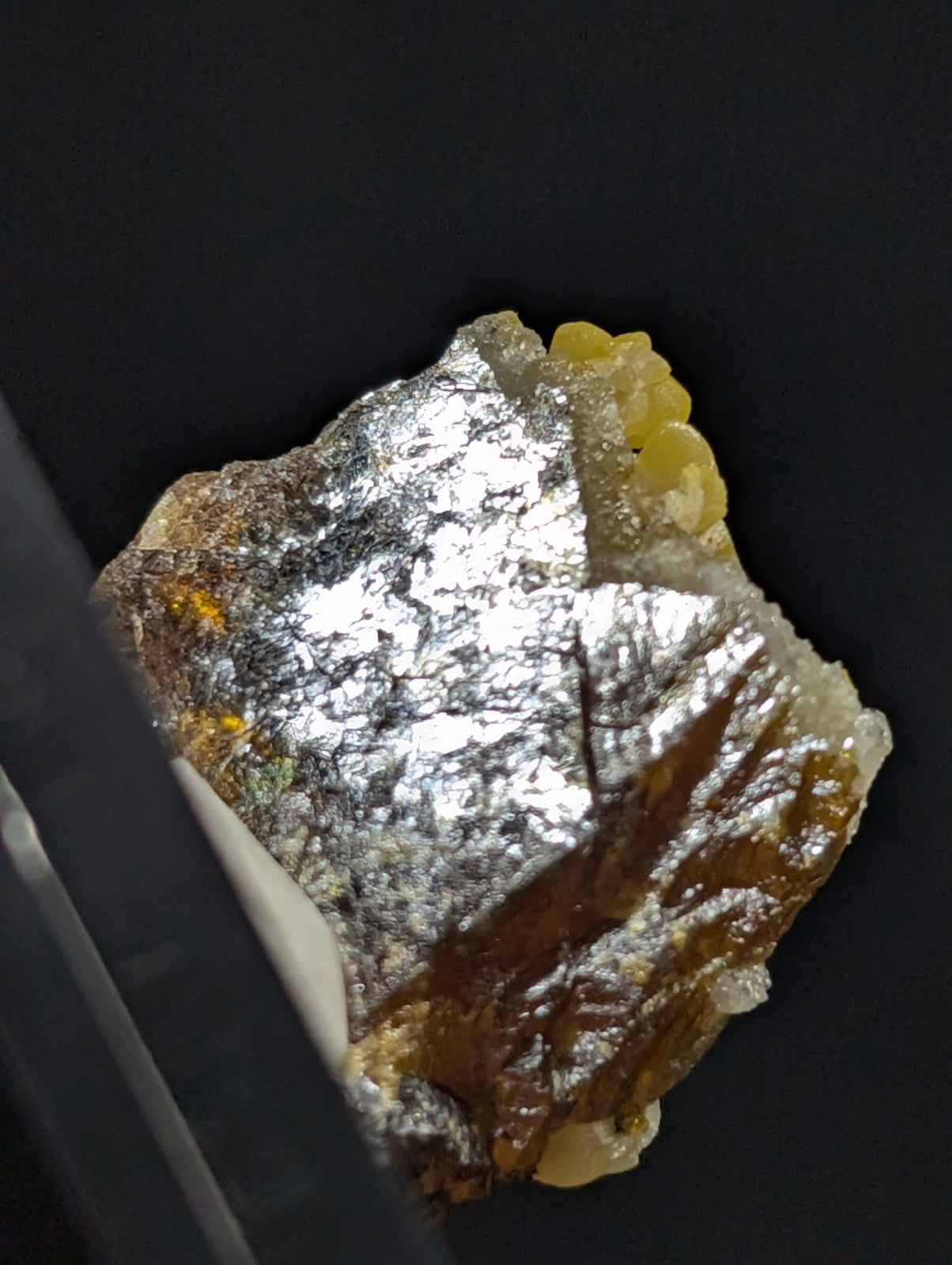 A metallic, irregularly shaped mineral specimen with a shiny silver surface and patches of yellowish-orange material.