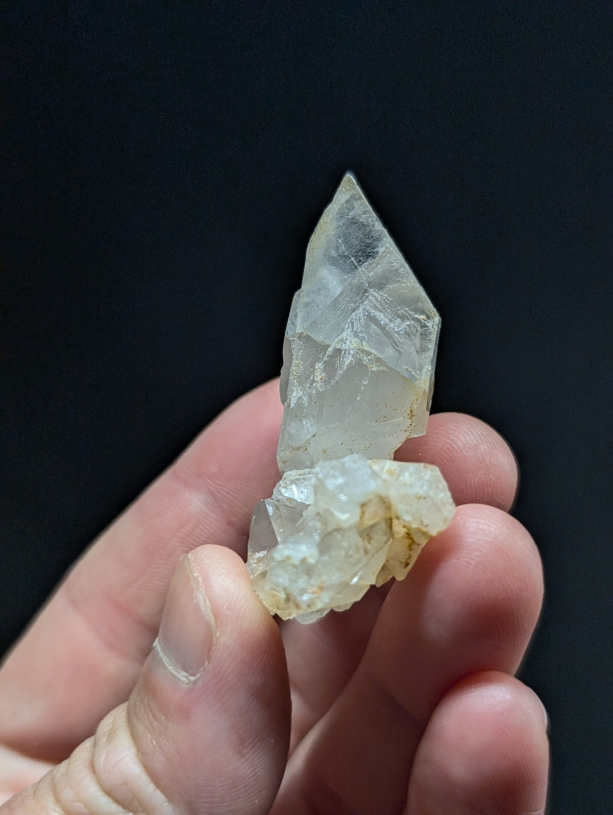 A translucent, pale quartz crystal with a pointed tip and rough, clustered base held gently in someone’s hand.