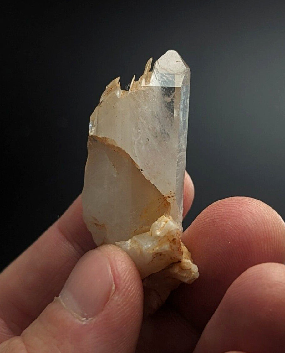 A translucent, pale quartz crystal with a pointed tip and rough, earthy base held gently in fingers.