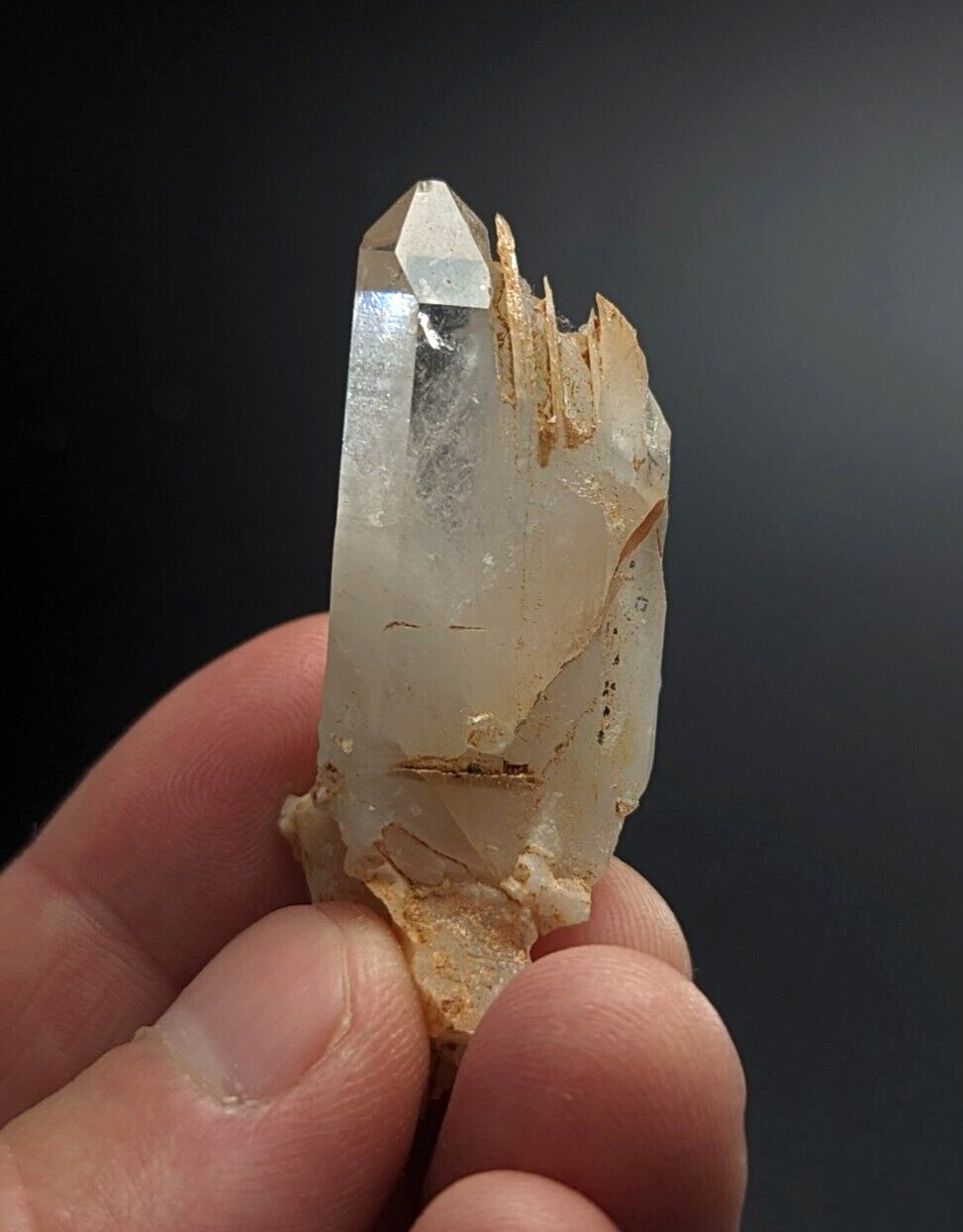 A translucent, clear quartz crystal with a pointed tip and natural brownish matrix at its base.