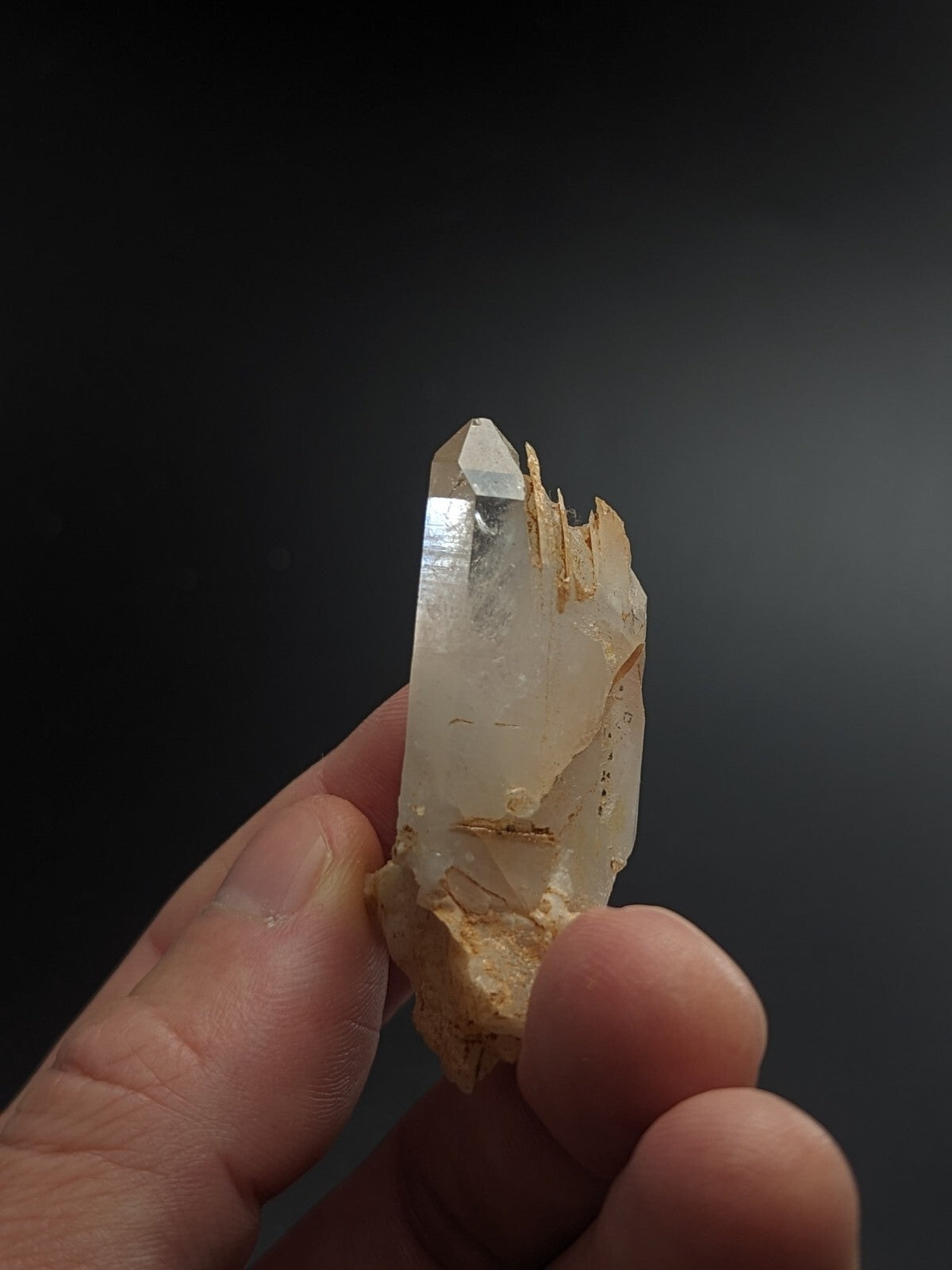 A translucent, pale quartz crystal with a pointed tip and rough, earthy base held gently in someone’s hand.