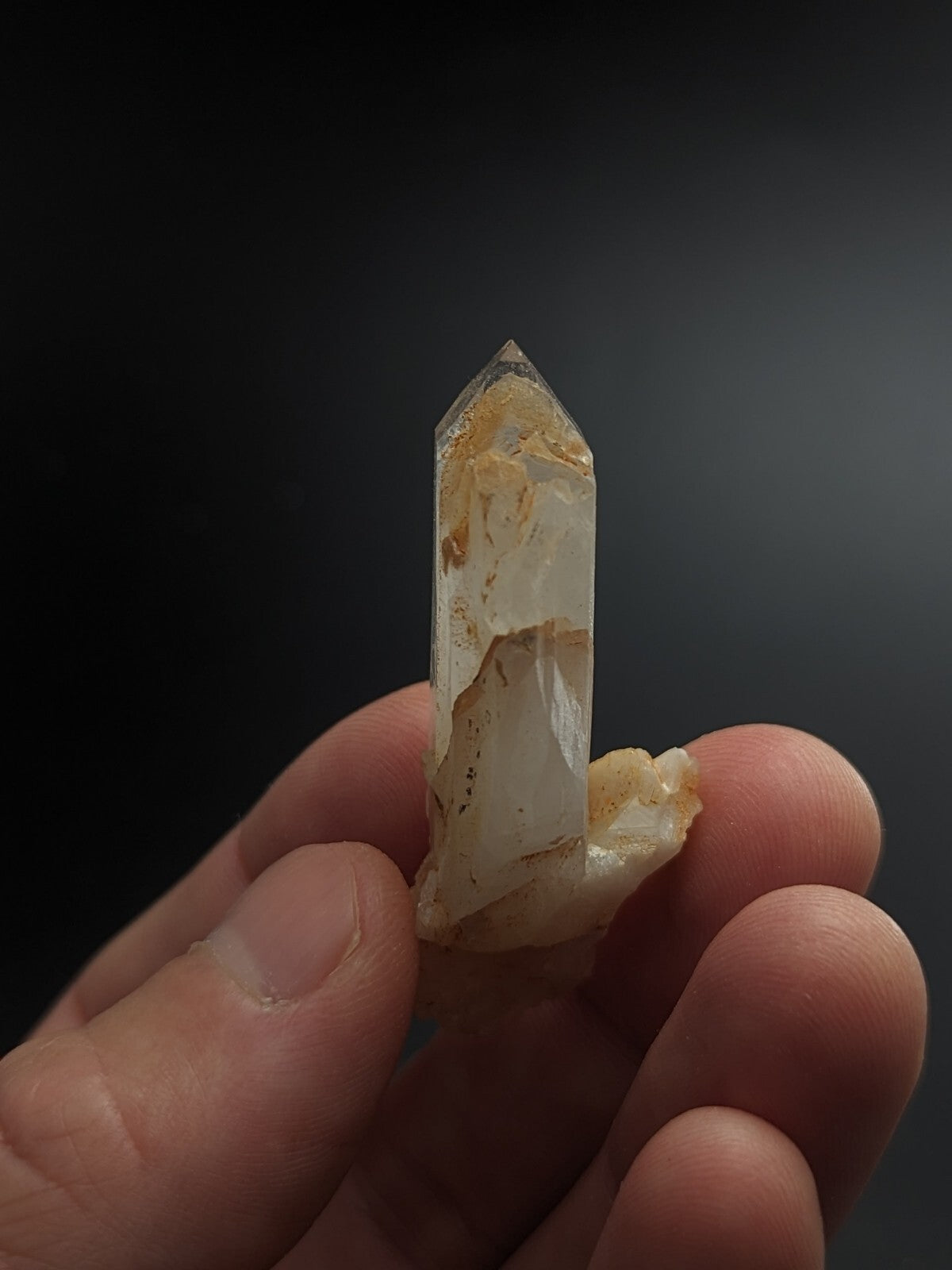A translucent quartz crystal with golden inclusions and a pointed tip, held gently between fingers.