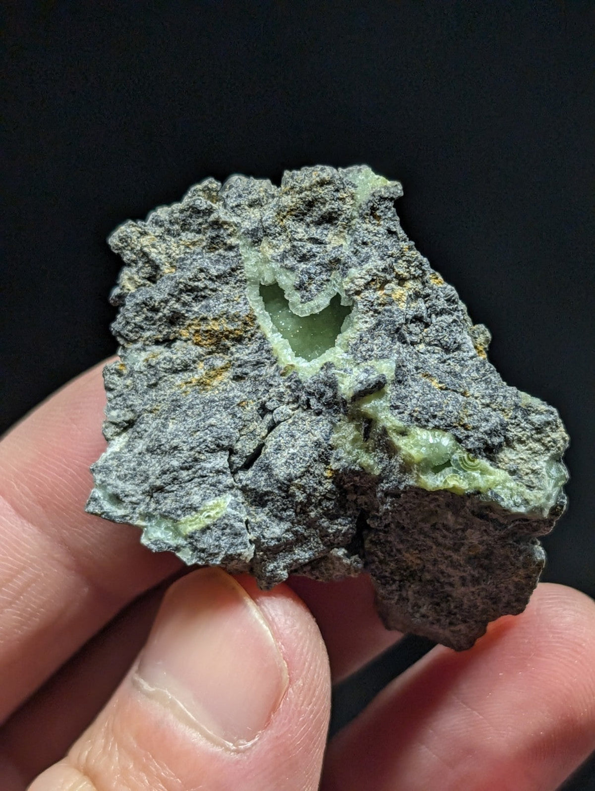 A rough, irregularly shaped piece of greenish-gray mineral with a visible hollow cavity and speckles of yellow.
