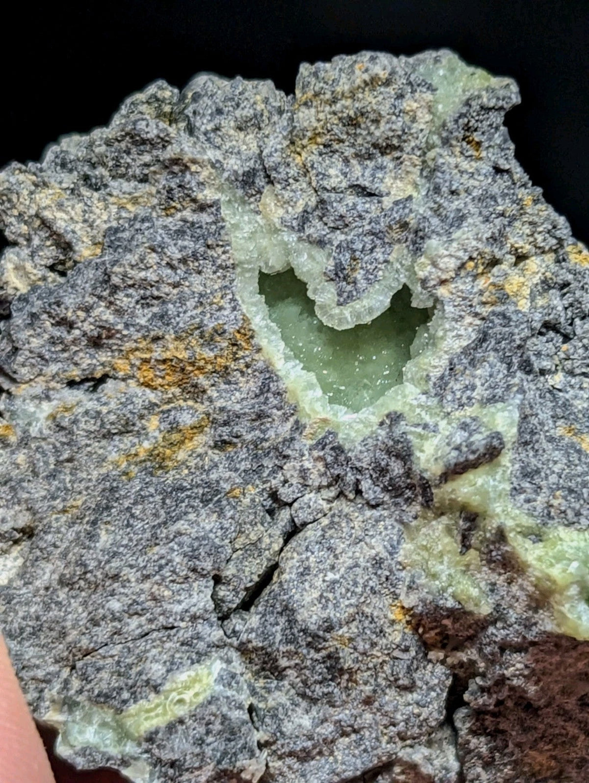 A heart-shaped cavity in a gray, speckled rock with greenish translucent crystals lining its edges and patches of yellow mineral streaks.
