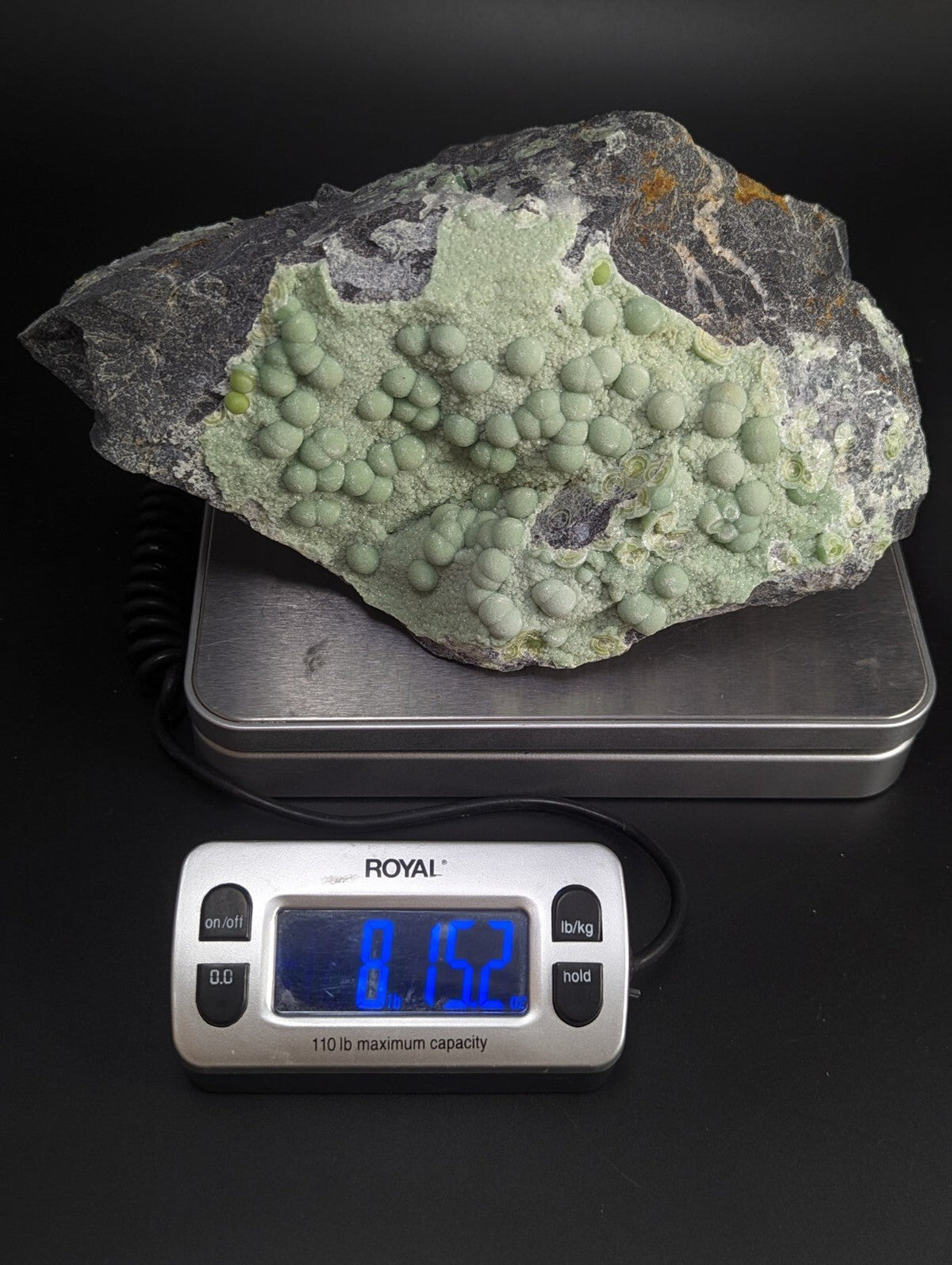 A greenish-gray mineral specimen with rounded, bulbous formations on a dark rock matrix.