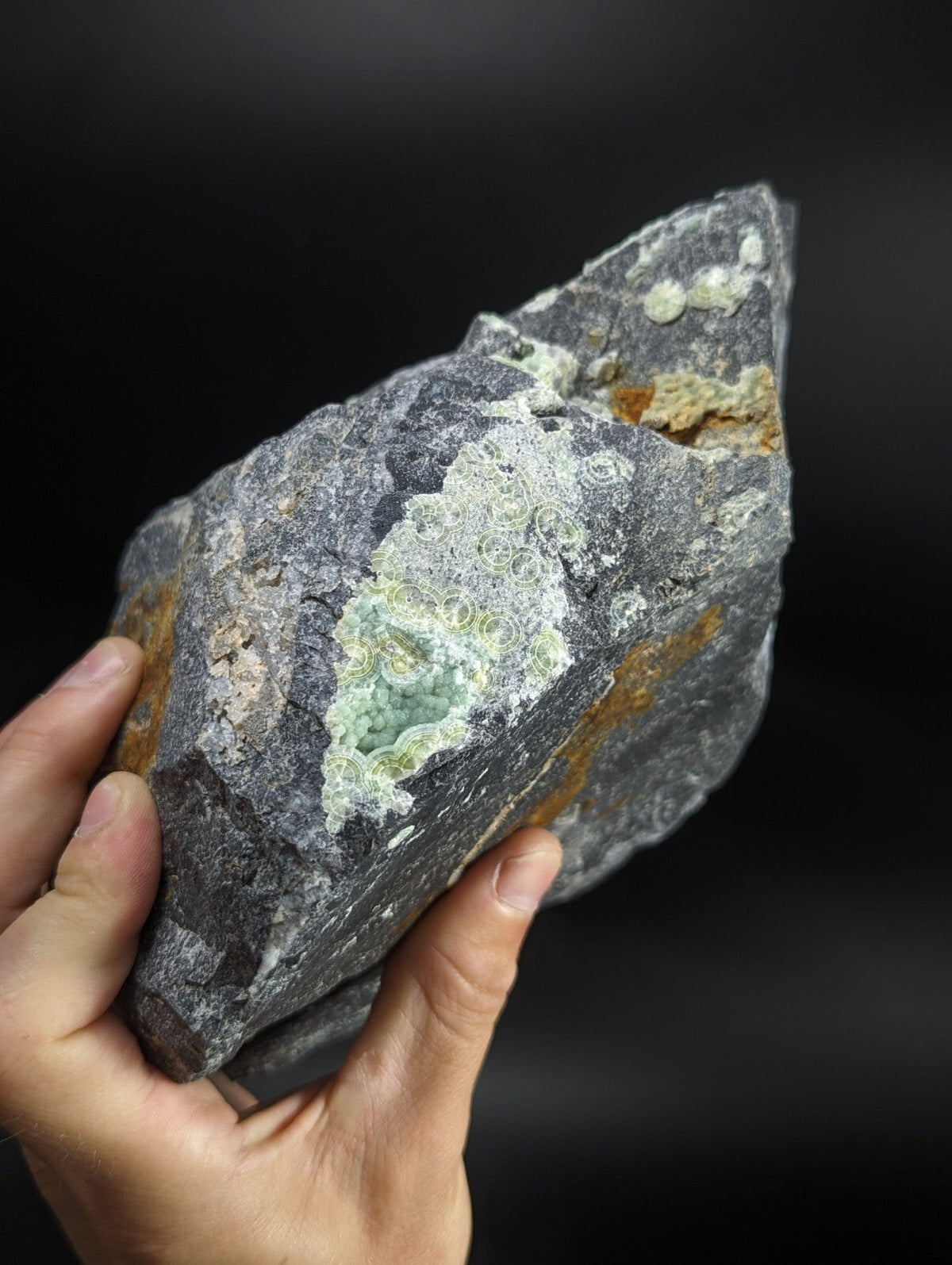 A rugged, dark gray rock with a striking patch of pale green mineral formations and hints of orange rust-colored streaks.