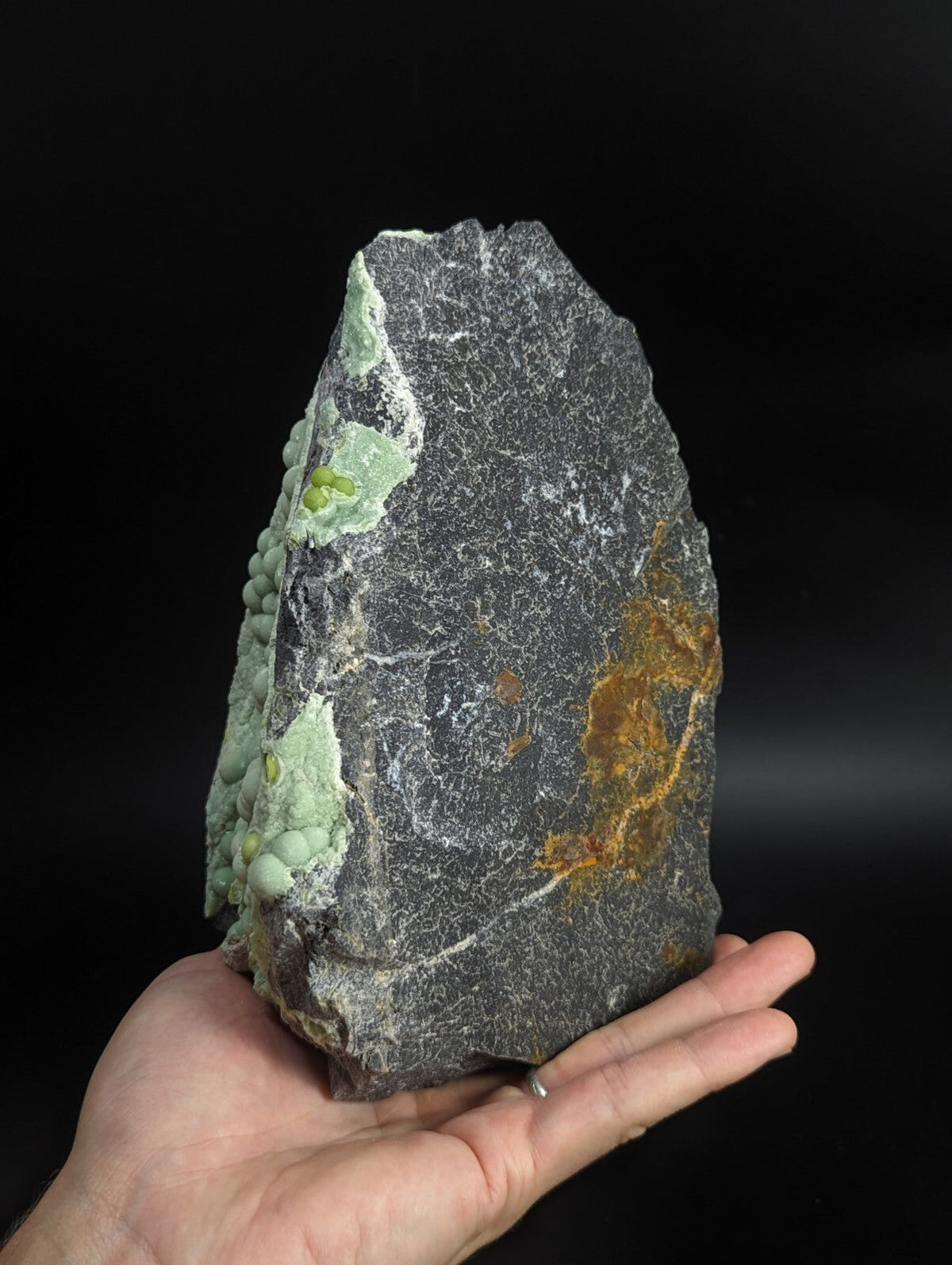 A hand holds a dark gray rock specimen featuring patches of light green mineral growth and streaks of orange-yellow oxidation.