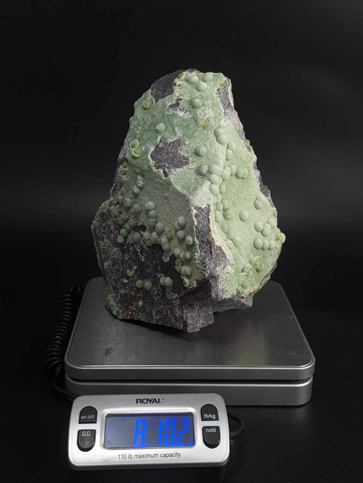 A green and gray mineral specimen with rounded, bulbous growths resting on a digital scale.