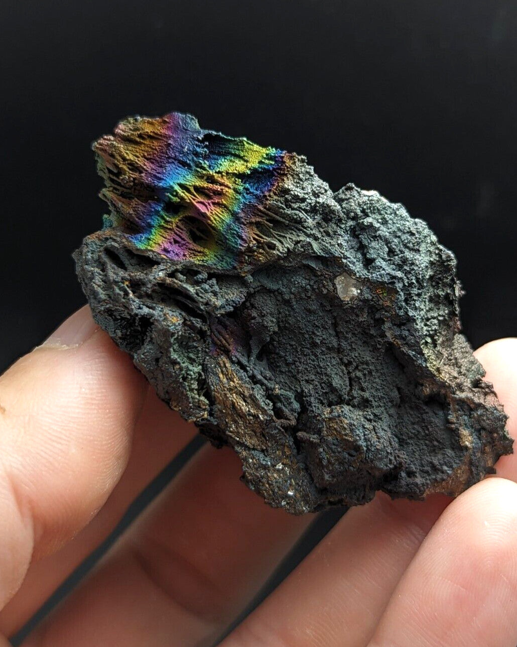 A hand holds a rough, dark gray rock with vibrant rainbow-colored mineral formations on its surface.