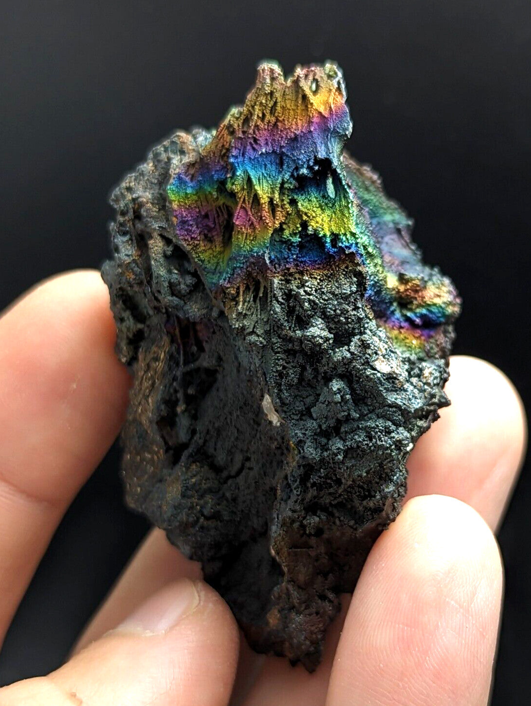 A hand holds a rugged, dark mineral specimen with iridescent rainbow-colored crystalline formations on its surface.