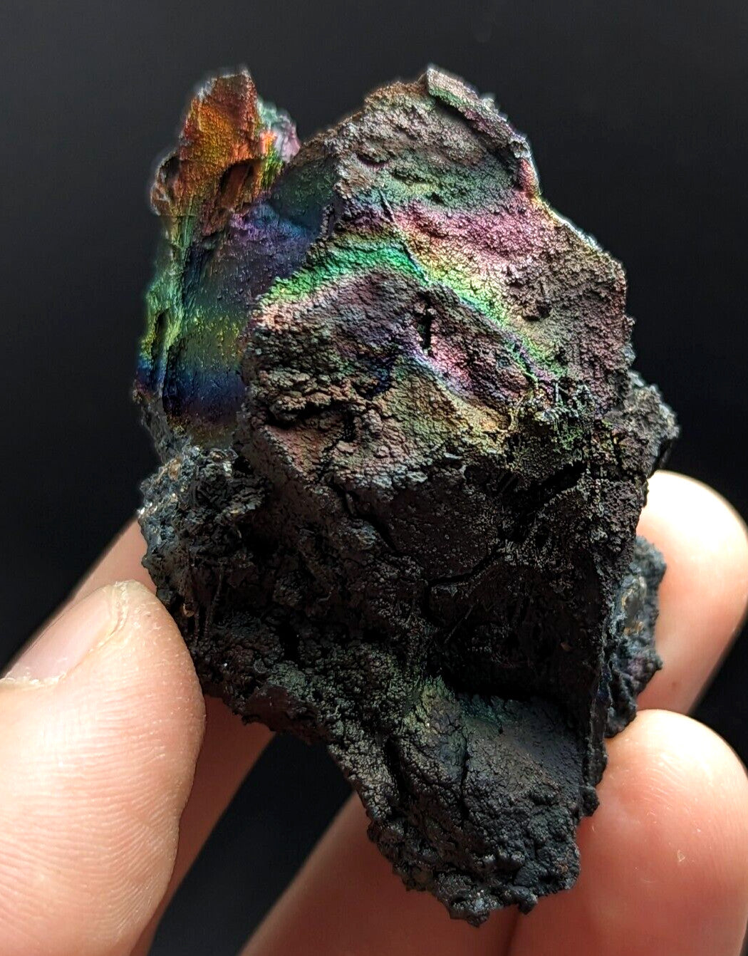 A rugged, dark mineral specimen with iridescent rainbow hues shimmering across its uneven surface.