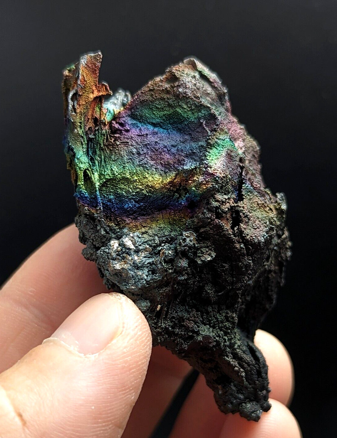 A rugged, irregularly shaped piece of corroded metal with a vibrant iridescent surface displaying rainbow hues across its textured, darkened form.