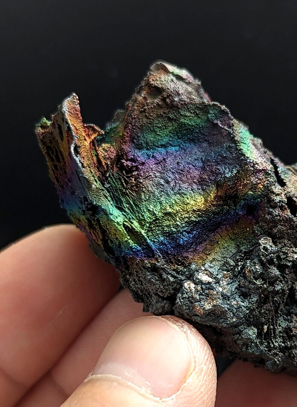 A hand holds a rough, irregularly shaped piece of iridescent metal with vibrant rainbow hues reflecting across its textured surface.