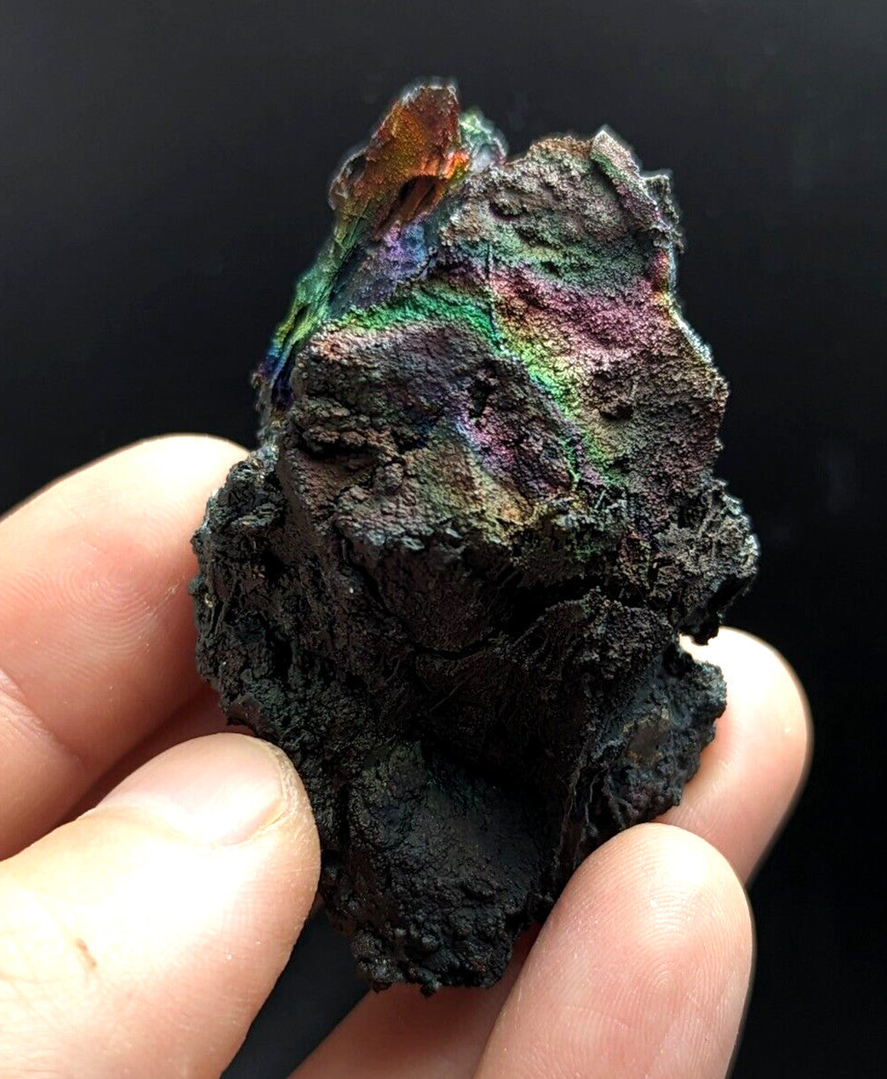 A hand holds a rough, dark mineral specimen with iridescent rainbow hues shimmering across its textured surface.