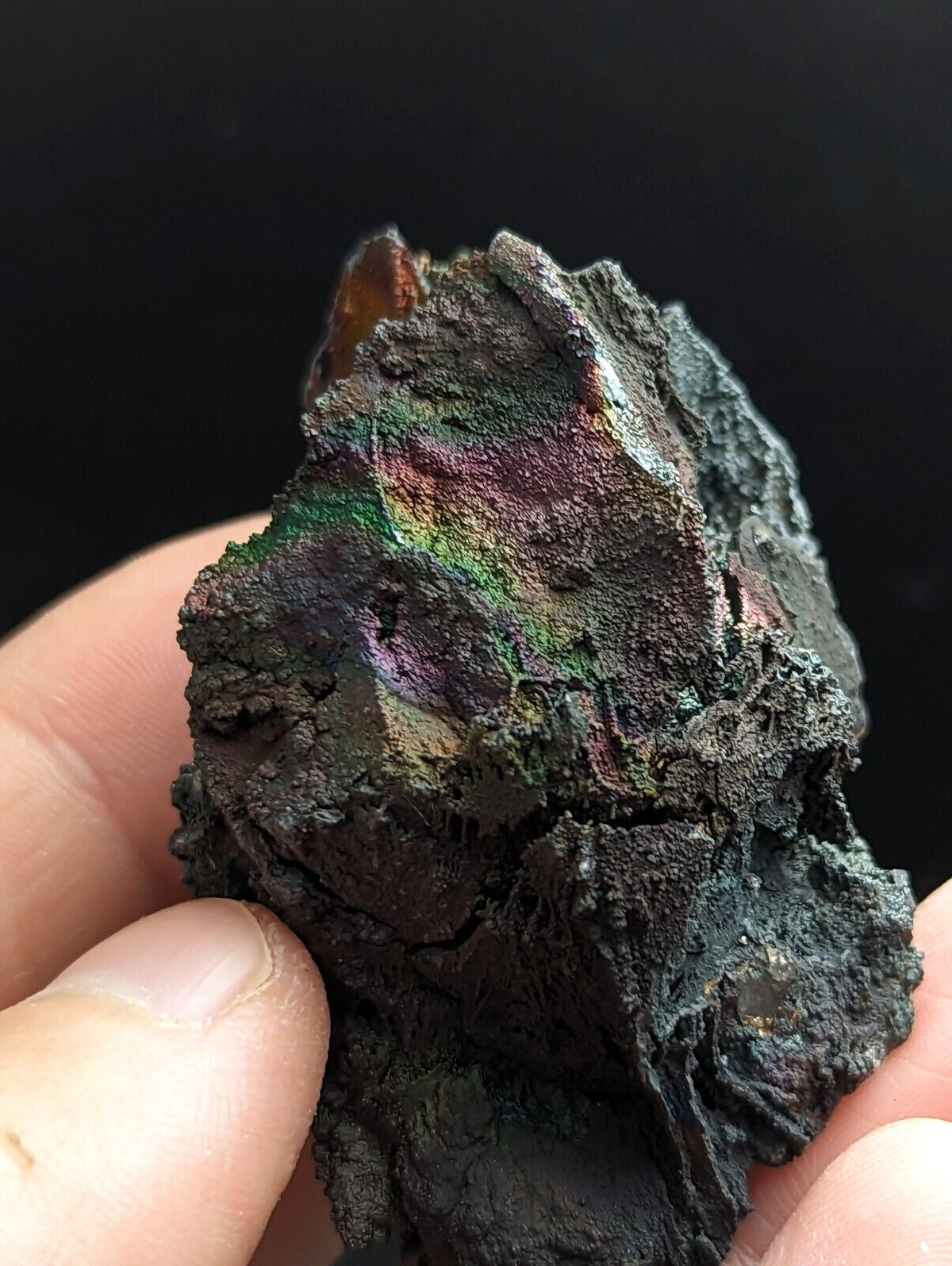 A rugged, dark metallic mineral specimen with iridescent rainbow hues of green and purple across its uneven surface.