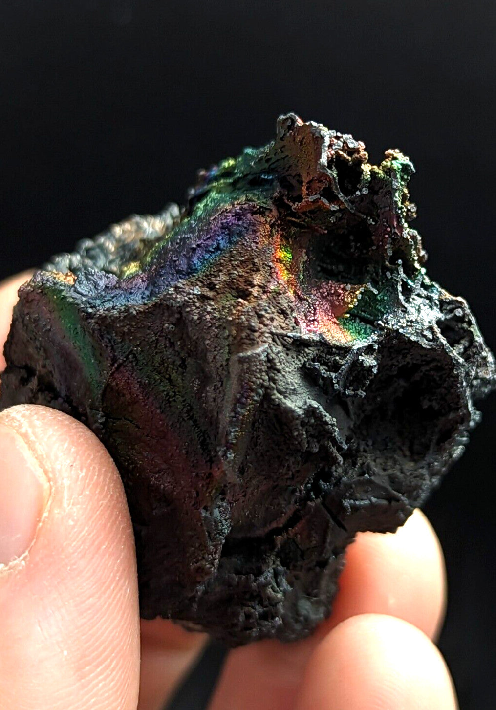 A rugged, dark metallic mineral specimen with iridescent rainbow hues shimmering across its uneven surface.