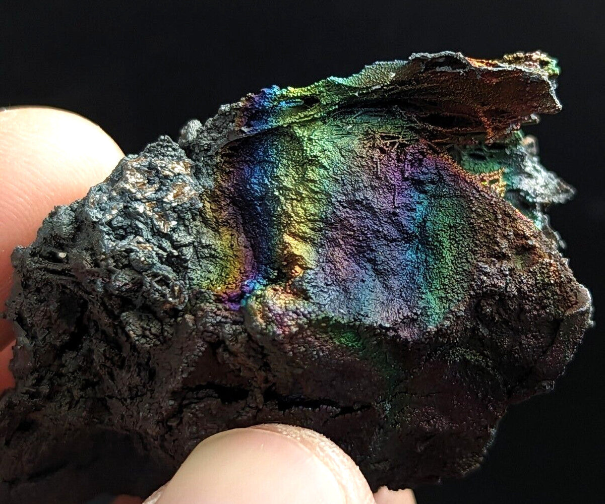 A rugged, dark metallic mineral specimen with a striking iridescent surface displaying vibrant rainbow hues of green, blue, purple, and gold.